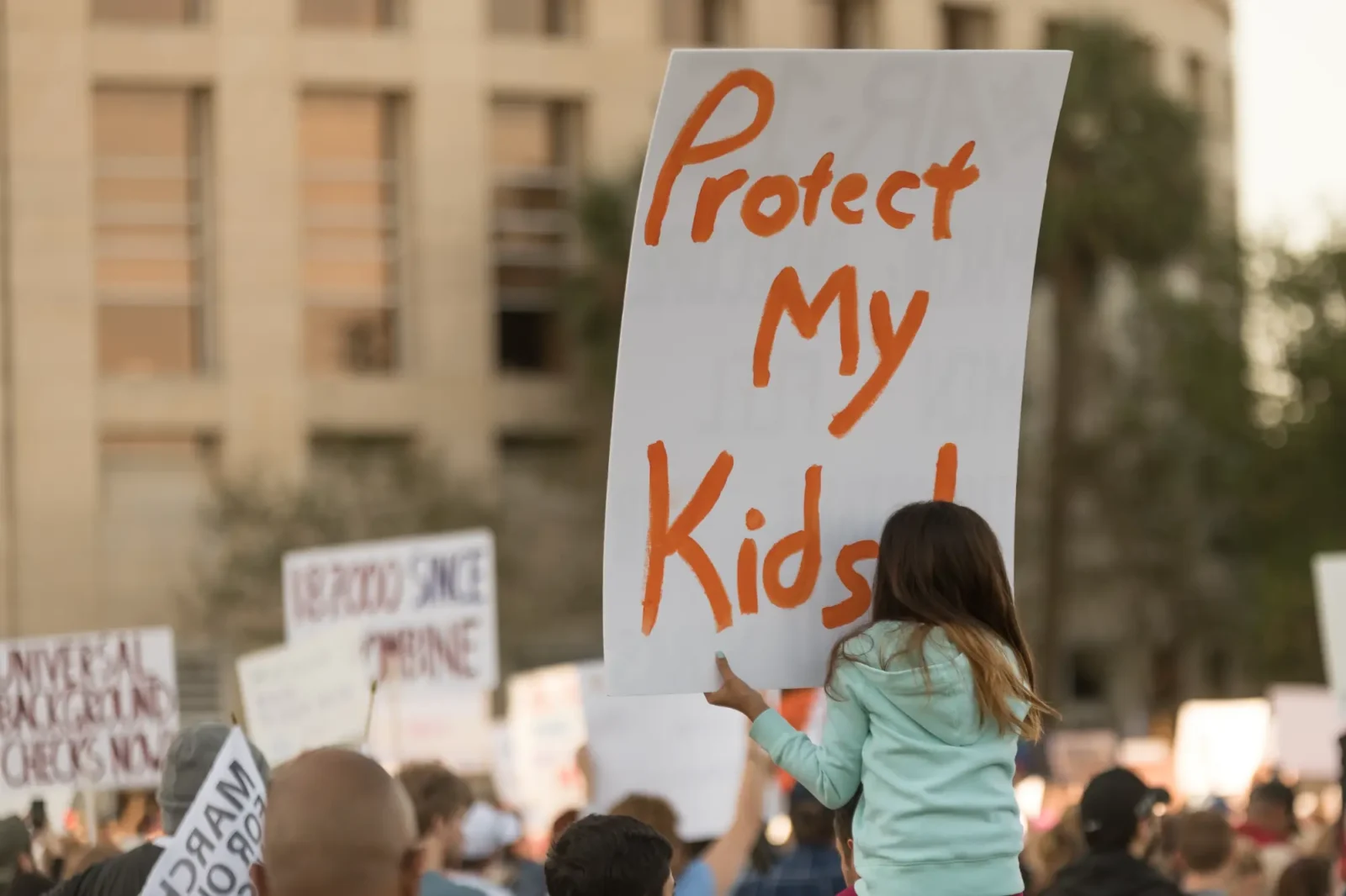 A young girl holds a sign reading "Protect My Kids!" high above a large crowd gathered for a public protest in an urban setting.