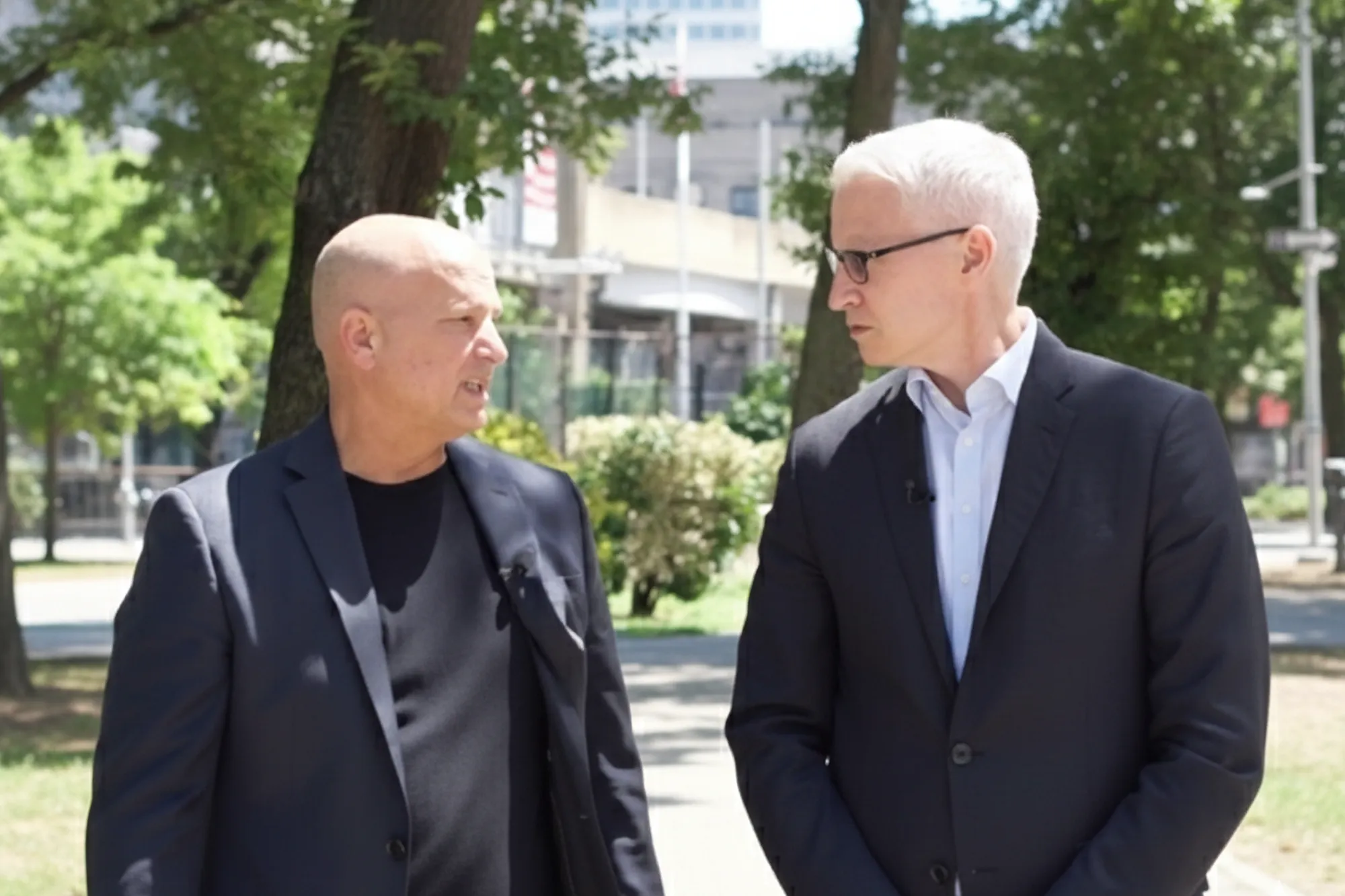 Doug Fierberg and Anderson Cooper, both in dark suits, look intently at each other while conversing in an urban park setting.