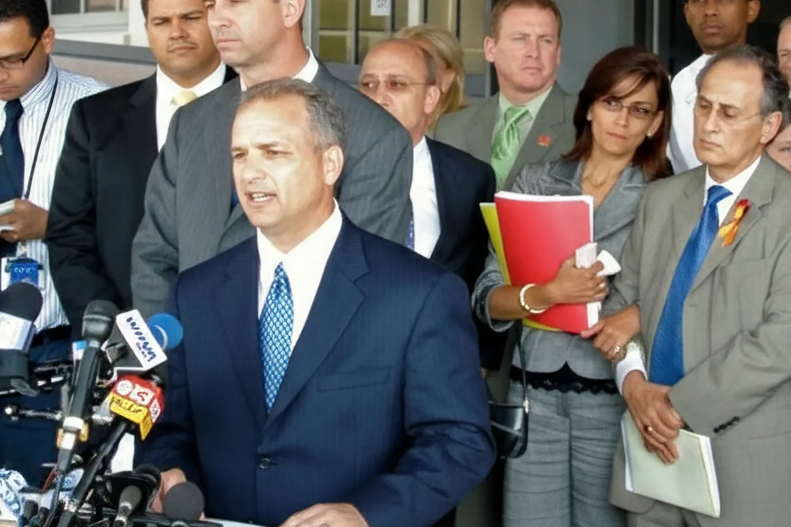 Attorney Doug Fierberg, wearing a blue suit, addresses reporters at a podium with several microphones. His serious colleagues stand behind him in a display of support.