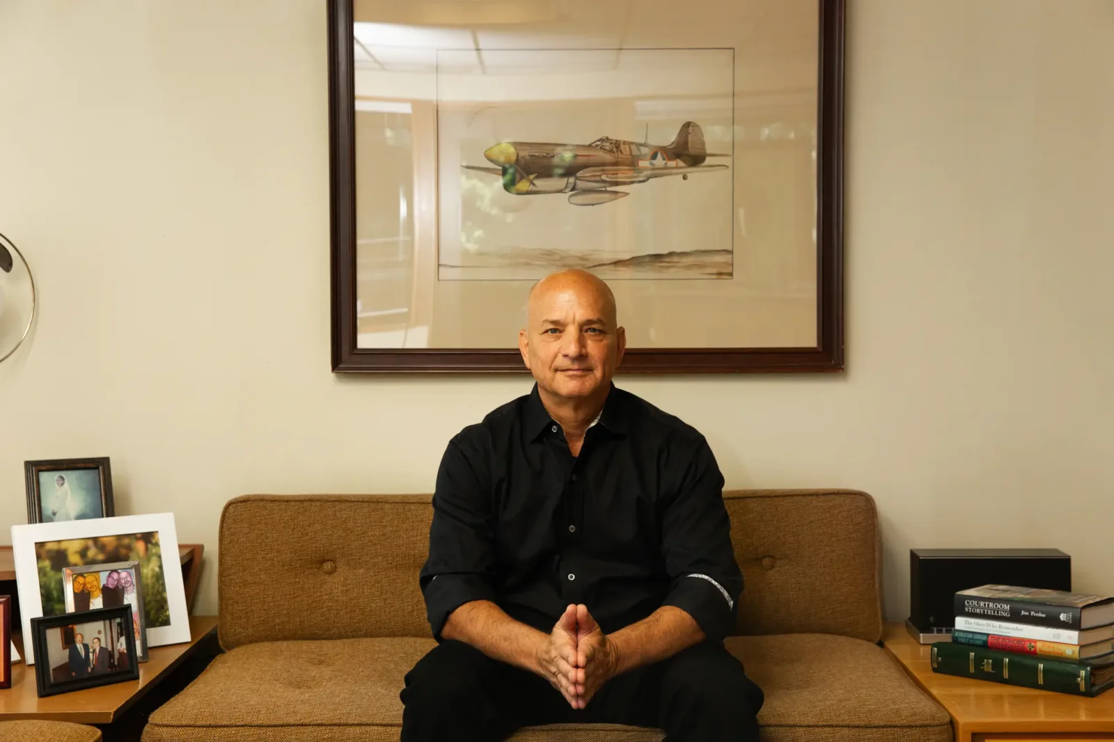 Doug Fierberg in a black shirt sits on a brown couch in an office, looking forward with his hands clasped. A framed painting of a military plane hangs above him.