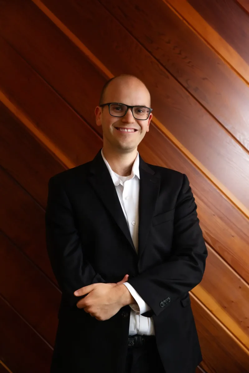 Jacob Goodman, a man wearing a black suit and glasses stands with a friendly smile, arms loosely crossed, against a wooden wall.