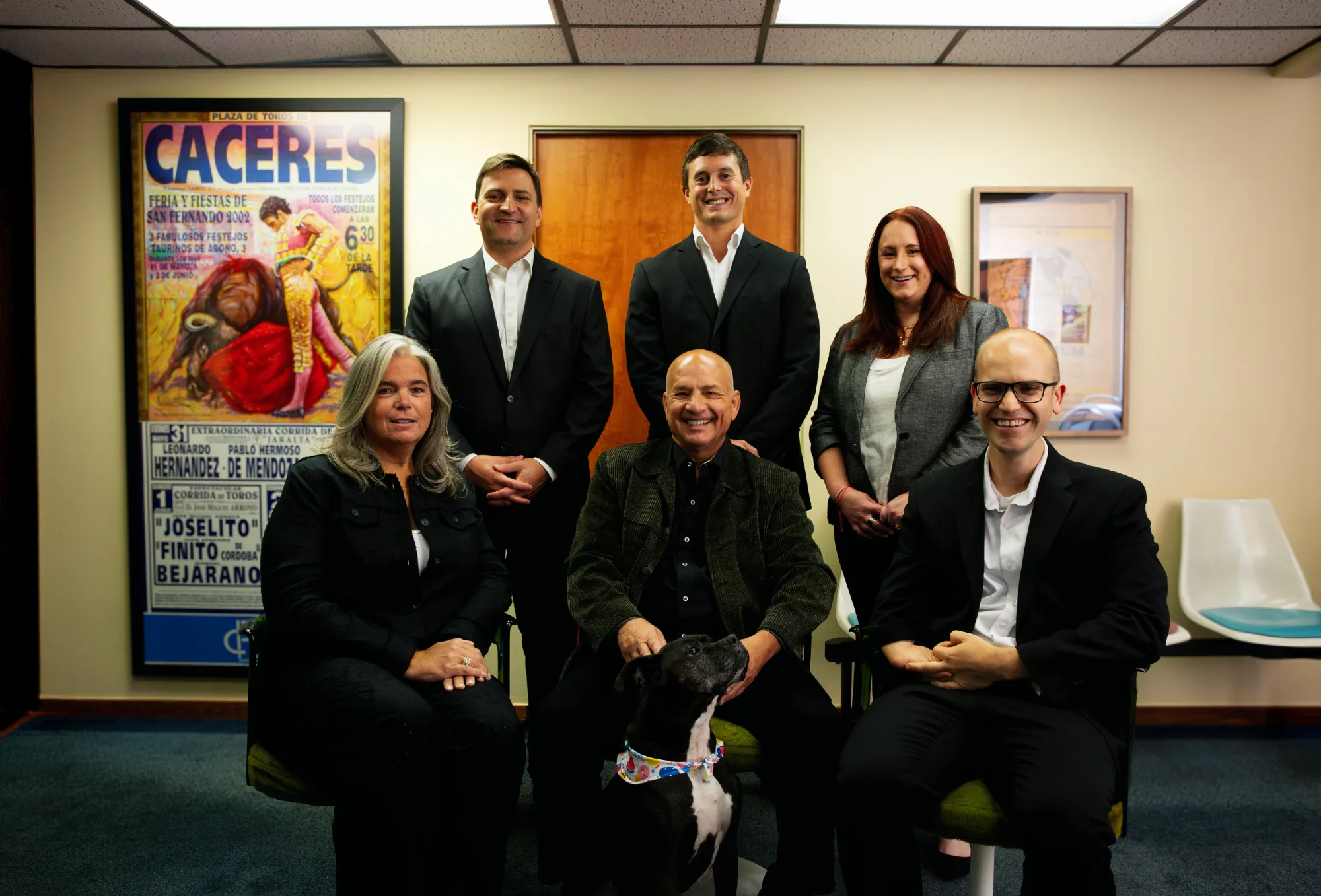 The Fierberg National Law Group team smile in a group portrait. The central seated man, Doug Fierberg, pets a friendly black and white dog wearing a colorful collar.