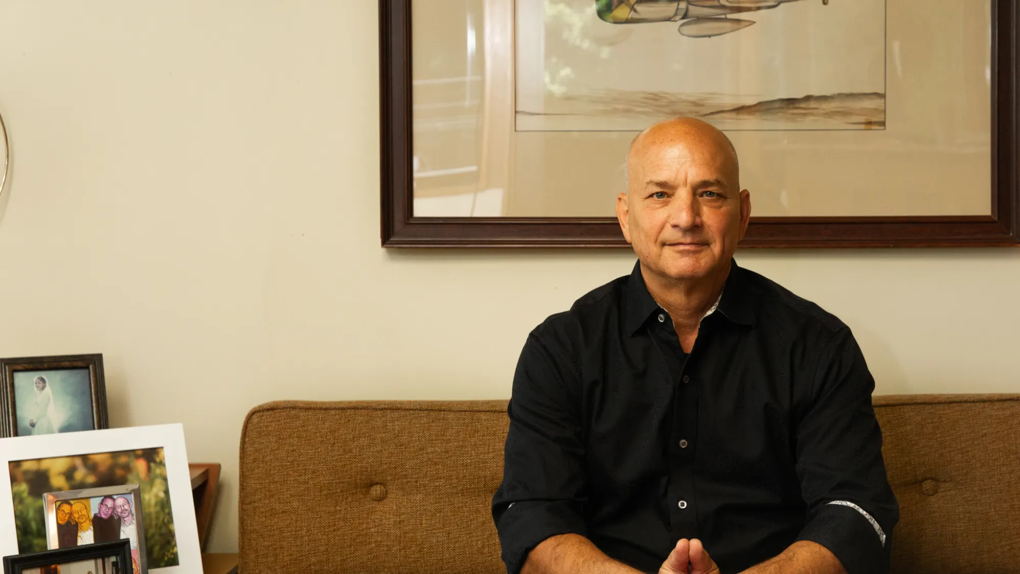 Doug Fierberg, a man wearing a black button-down shirt sits on a brown sofa. He looks straight ahead with a calm, professional expression in an office setting.