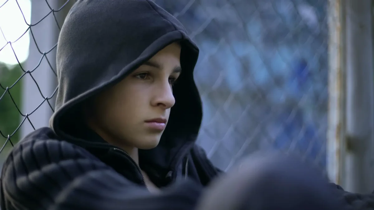 A young person wearing a dark hooded sweatshirt sits near a chain-link fence, looking down with a serious and pensive expression.