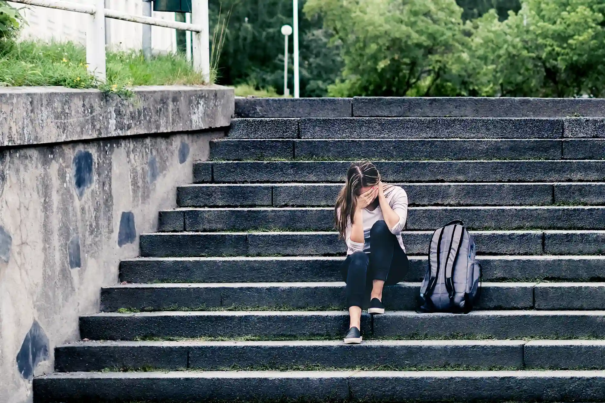 A young person with long hair sits on wide stone steps outdoors, their face covered by their hands in apparent distress. A gray backpack is nearby.