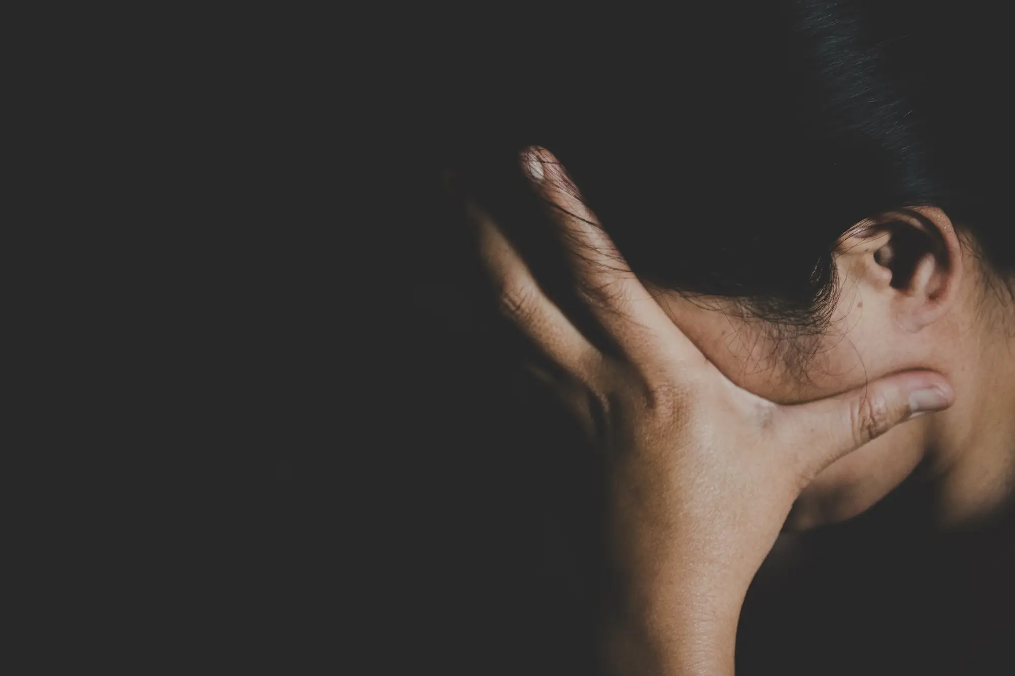 A close-up in low light shows a person pressing their hand against the side of their head in a pose of distress.