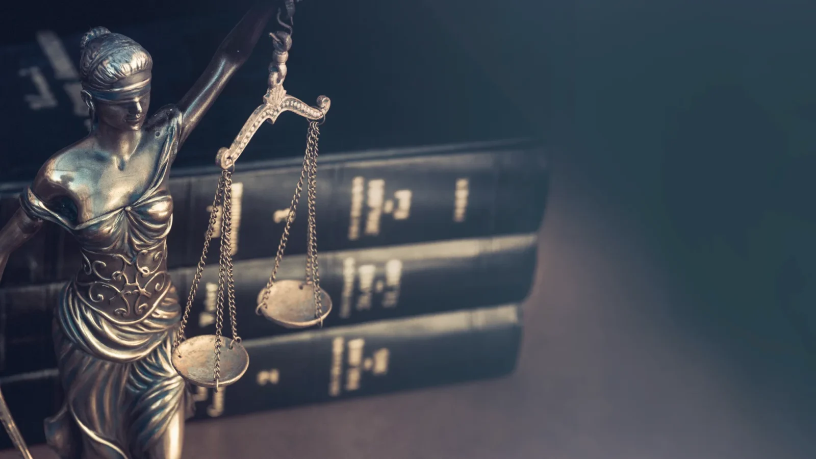 A bronze statue of Lady Justice, blindfolded and holding the scales of justice, with a stack of dark law books behind her.