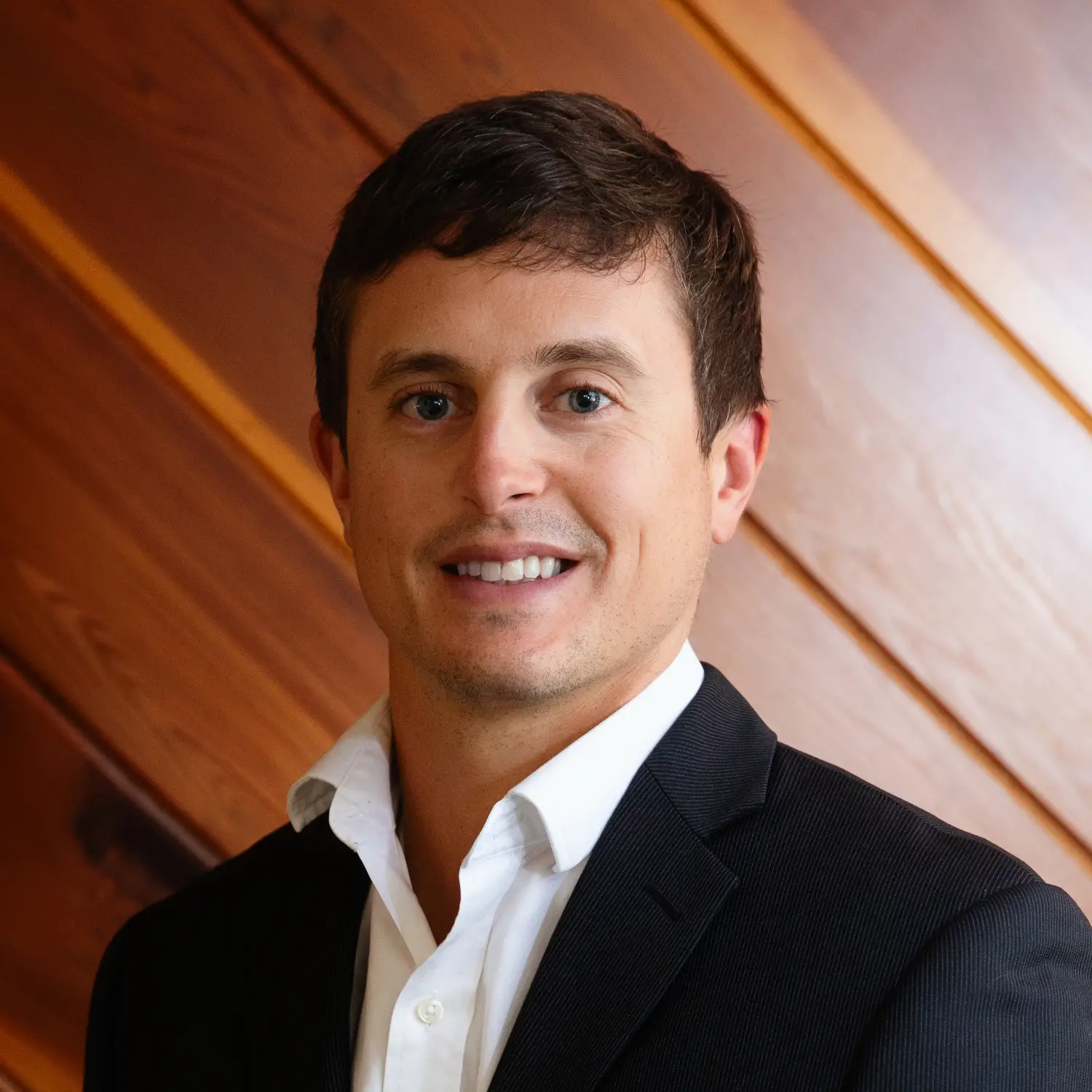 A headshot of Bailor Bell, smiling brightly while wearing a black suit jacket and white open-collar shirt against a wood wall.
