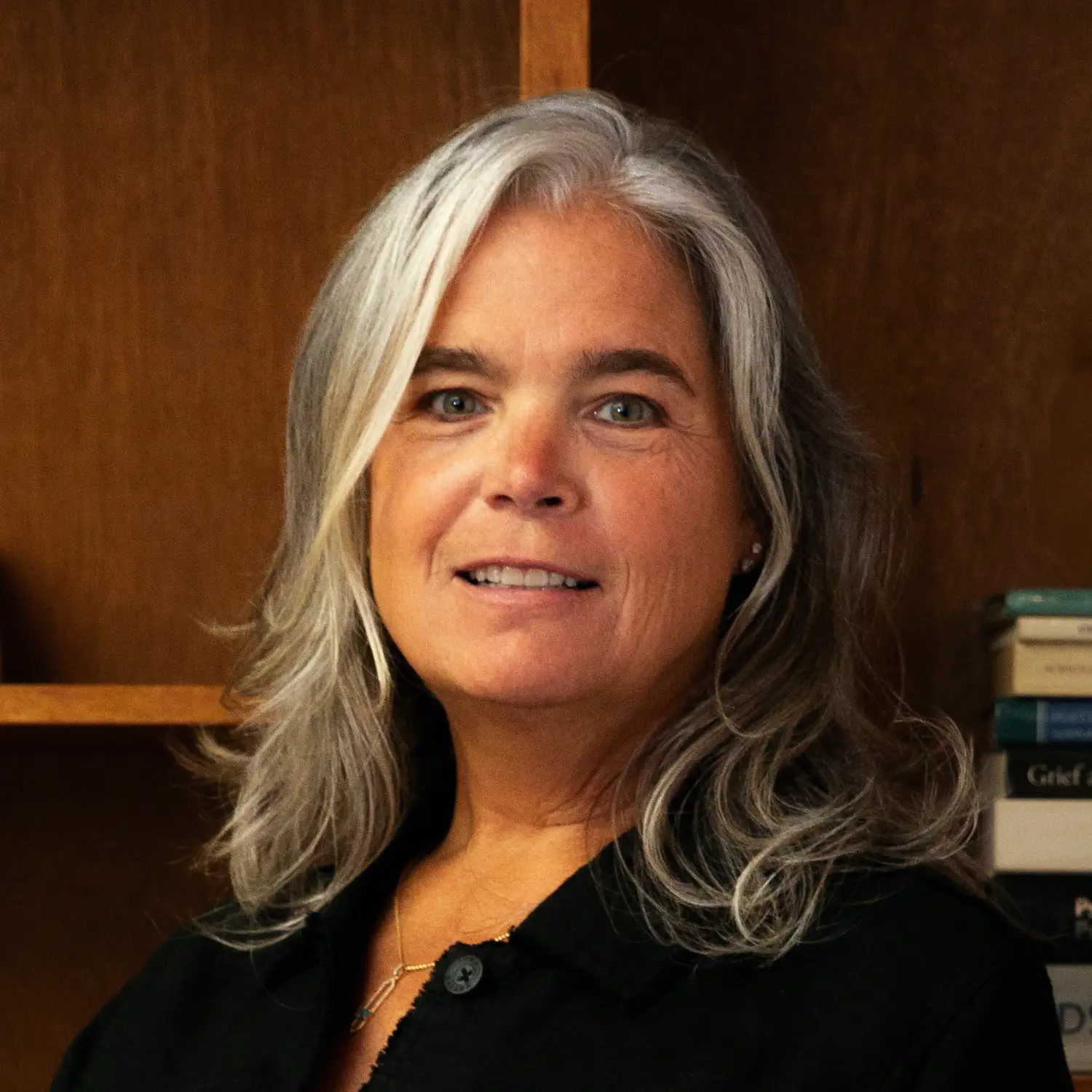 Chris Dejong smiles warmly. She has long gray hair and wears a dark shirt against the backdrop of a wooden bookshelf.
