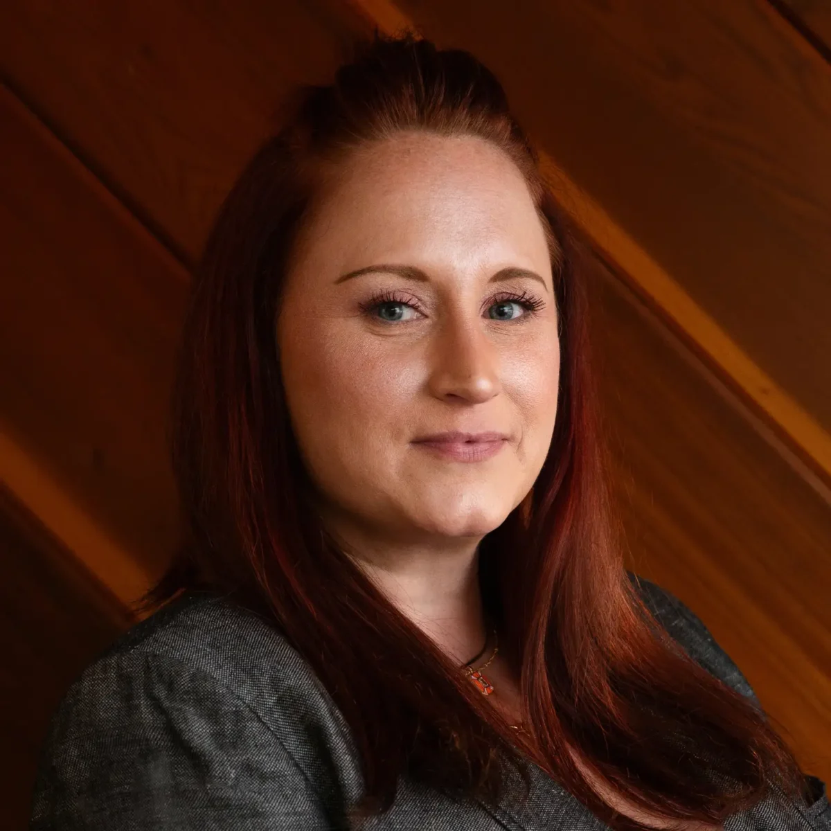 Erika Klockziem, with red hair and a slight smile, wears a dark gray blazer for this professional headshot against a wooden wall.