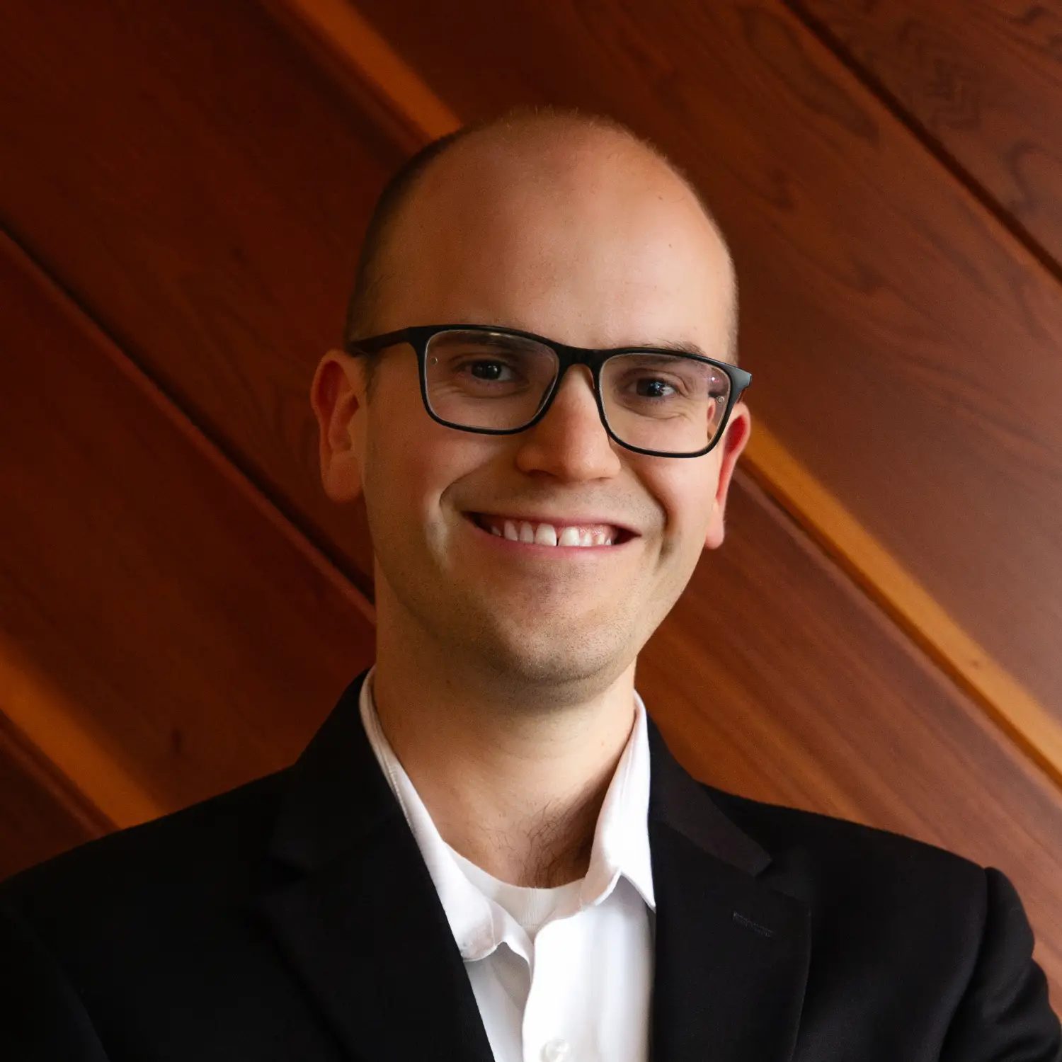 Jacob Goodman, wearing glasses and a black suit, smiles widely for a professional portrait against a warm wooden background.
