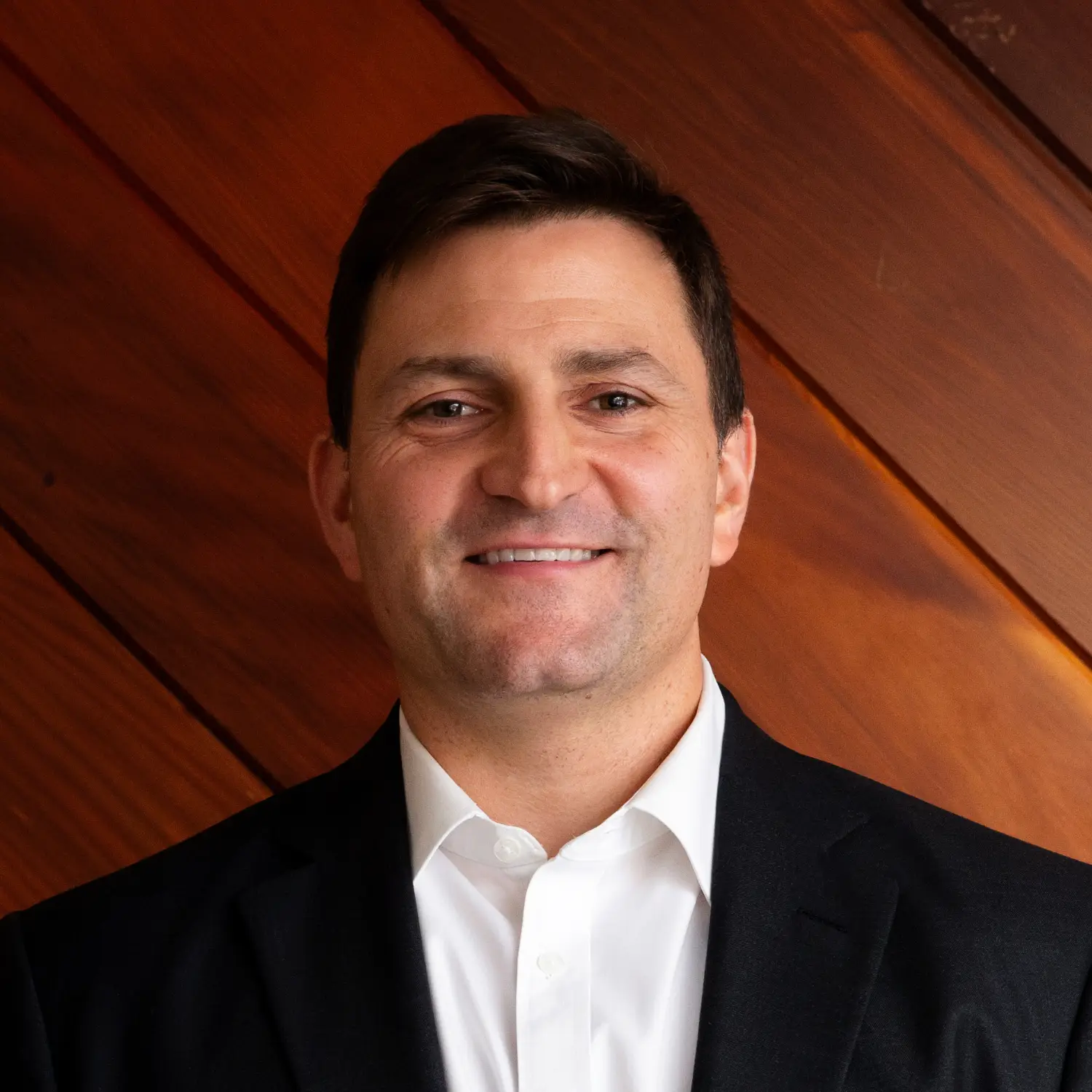 Jonathon Fazzola, wearing a black suit and white shirt, stands against a warm wood-paneled wall, looking confidently at the camera with a friendly smile.