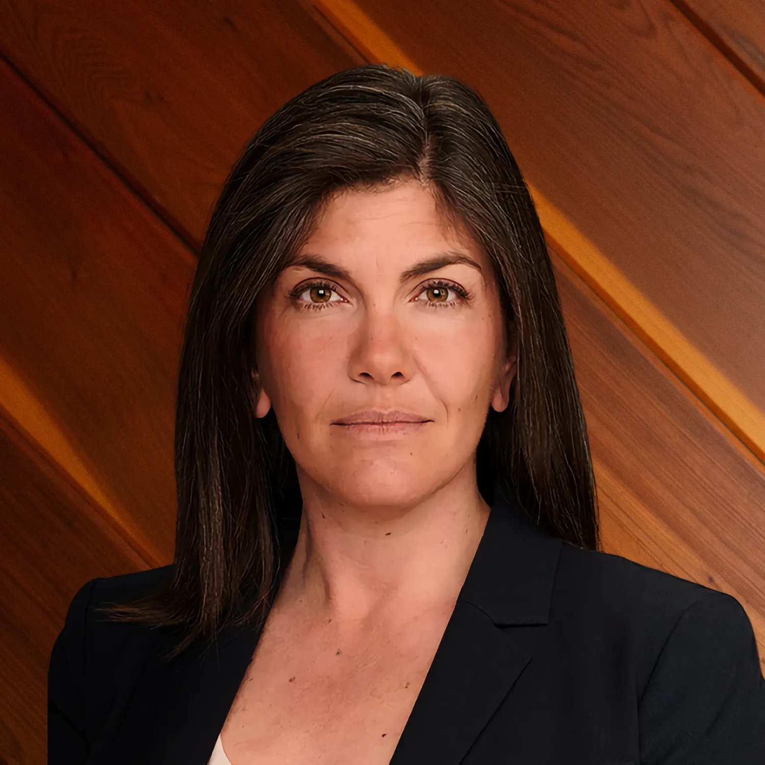 A professional headshot of Cari Simon, a woman with long brown hair, wearing a black blazer and looking directly forward against a background of warm wooden panels.