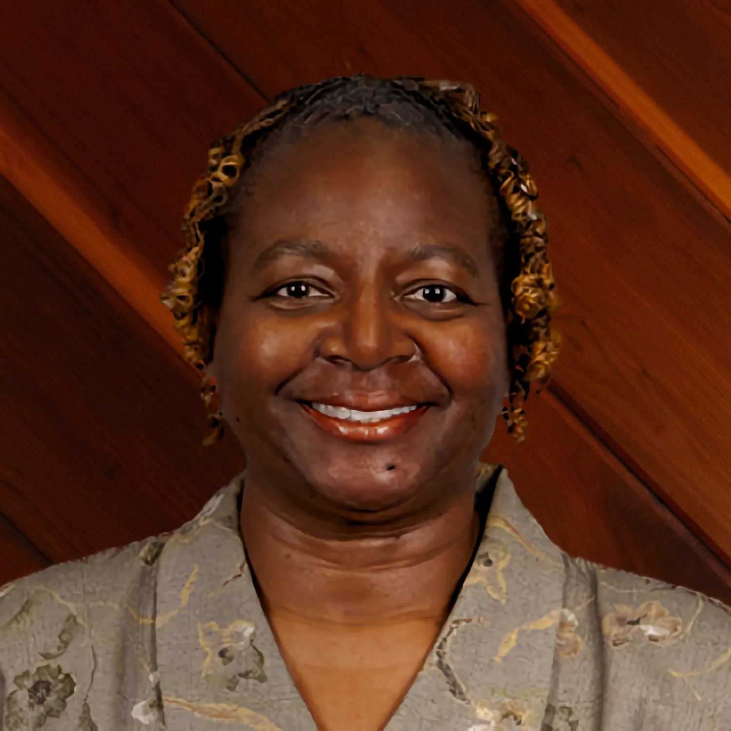 Darcell McCoy smiles warmly in a professional headshot. She has curly, highlighted hair and wears a patterned gray blouse, standing against a dark wood-paneled background.