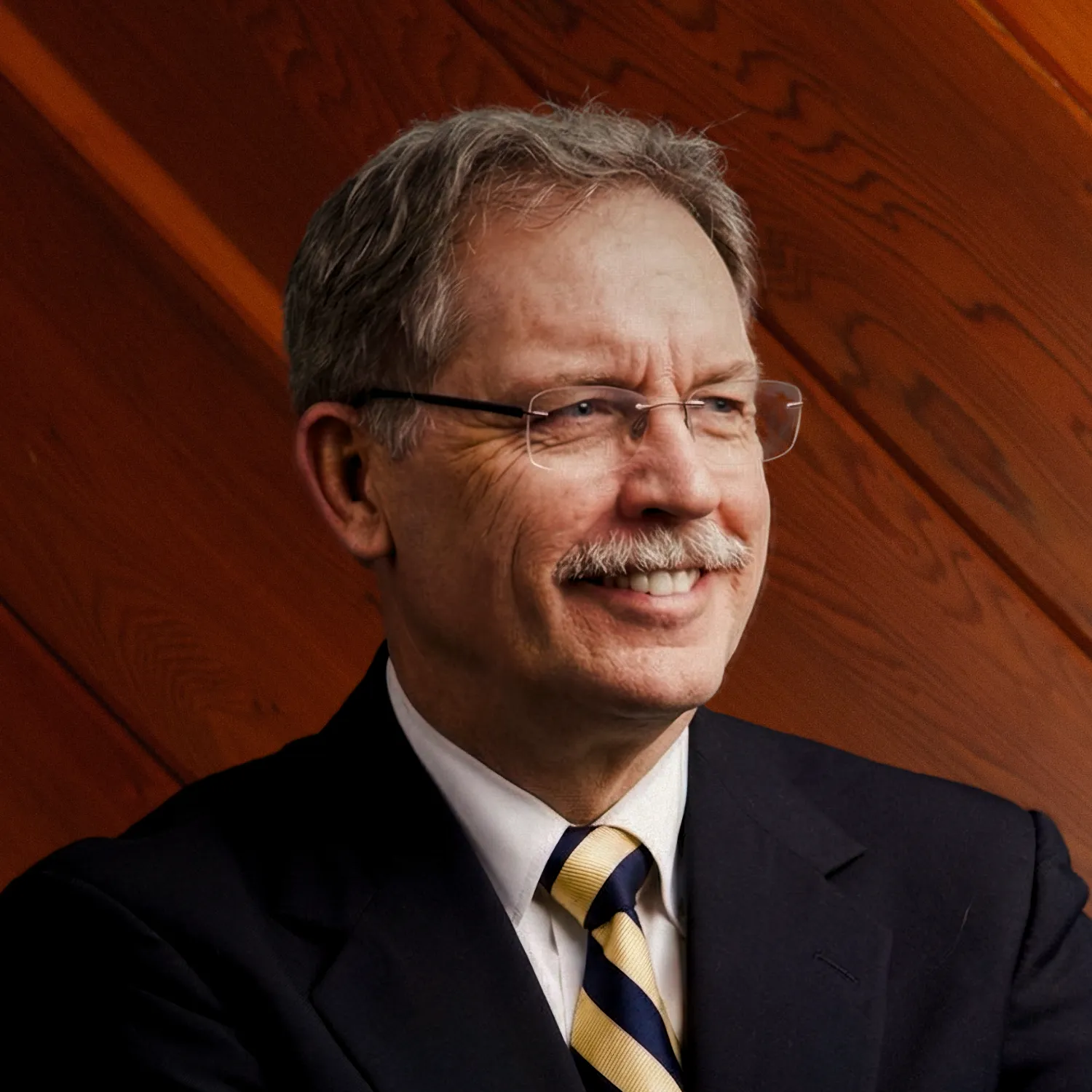 George Thompson wears a dark suit, striped tie, and glasses, smiling confidently against a background of wooden paneling.