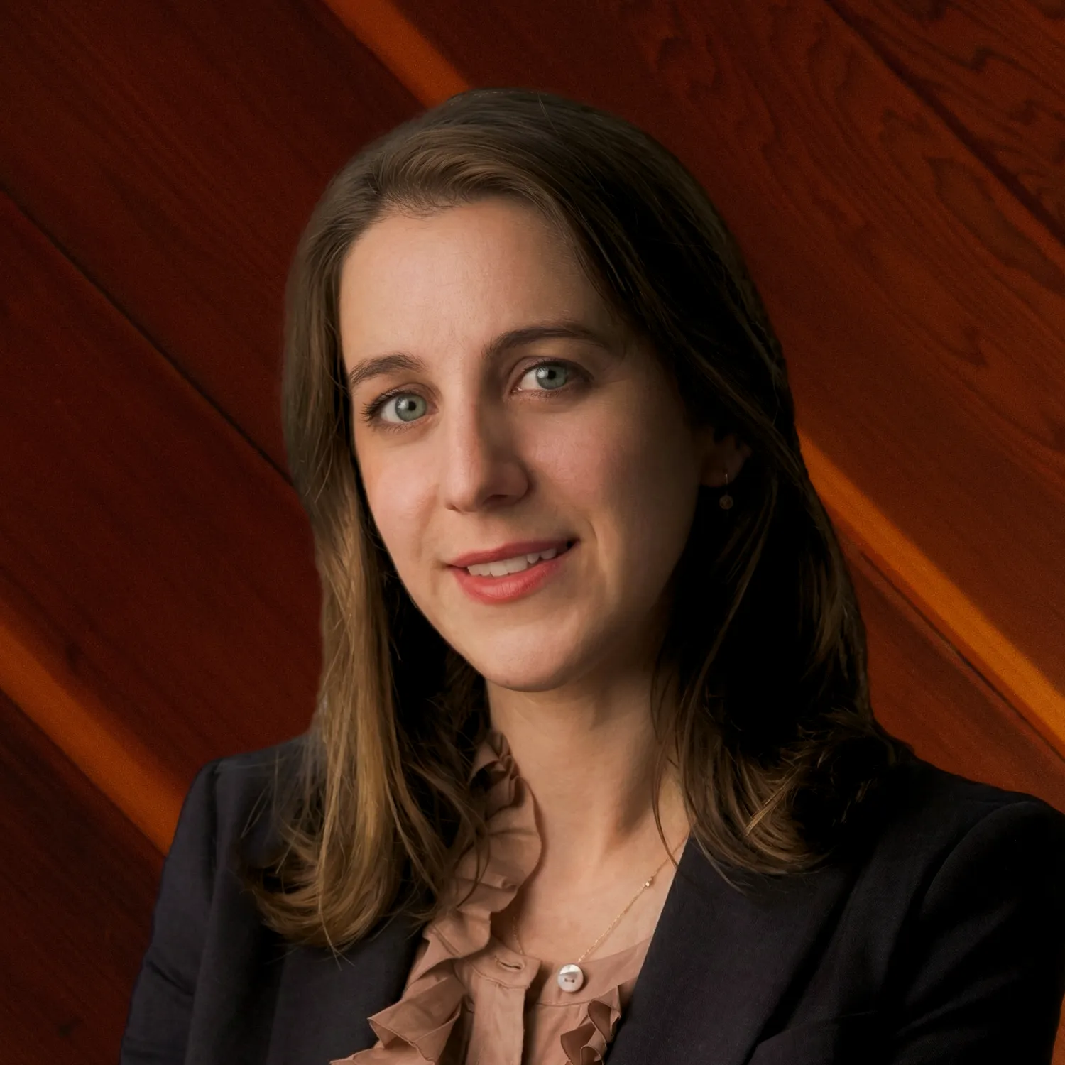 Olympias Iliana Konidaris, an attorney, smiles in a professional headshot. She wears a dark blazer over a ruffled blouse against a warm wood paneled background.