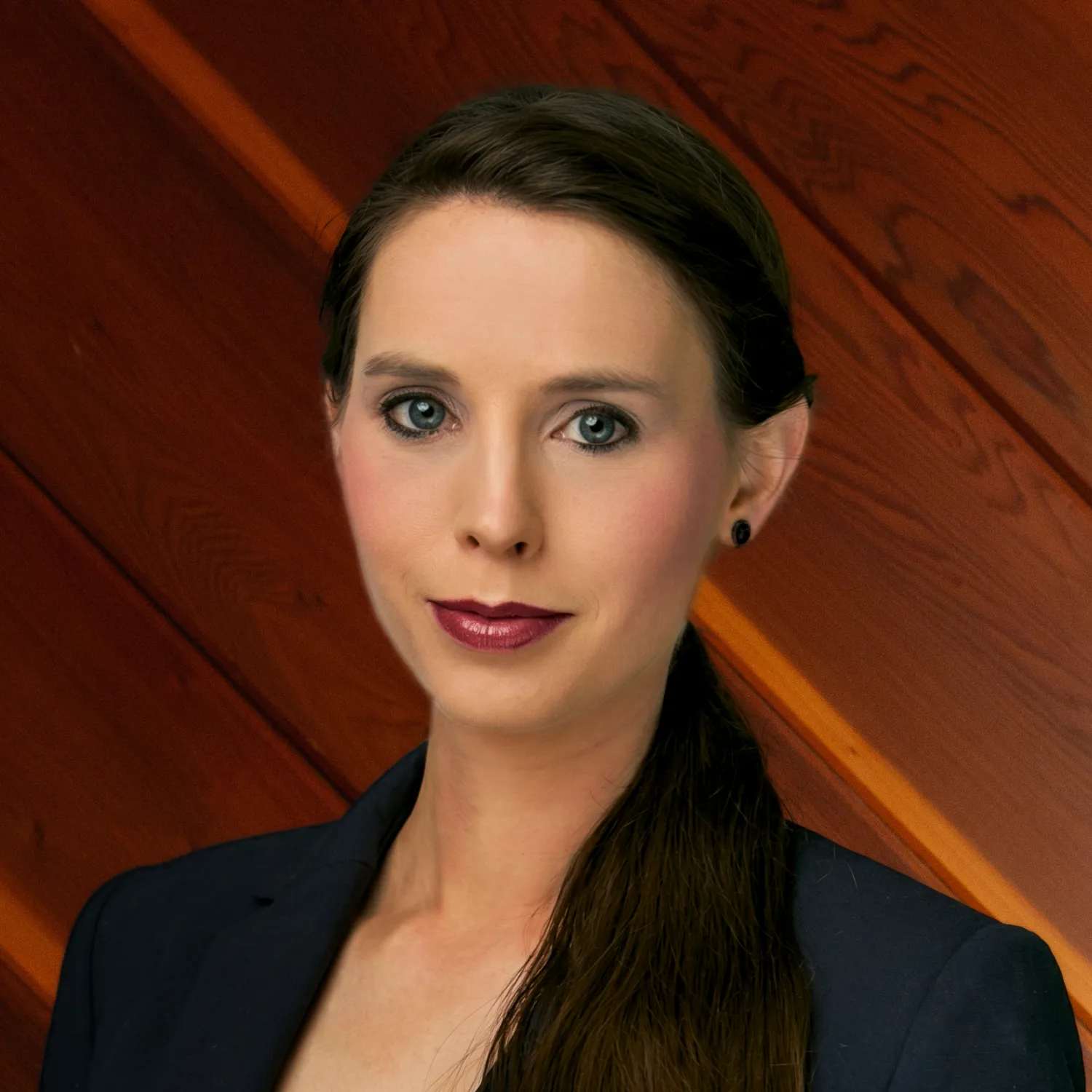 Rachael Denhollander in a dark business blazer, looking forward with a neutral, professional expression. Her dark hair is styled in a sleek ponytail.