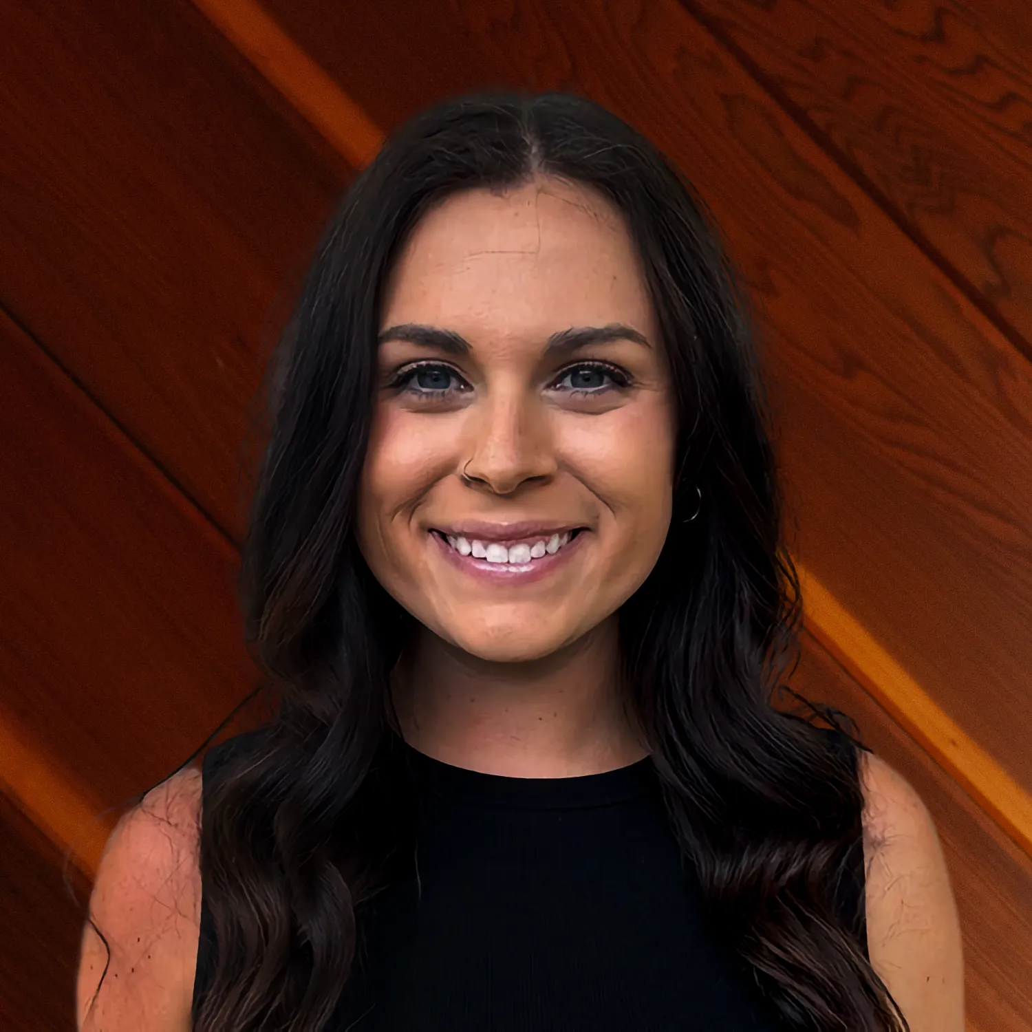 Weslie Caprai, wearing a black top, smiles warmly for a headshot. She has long dark hair and stands against a diagonal wooden wall.