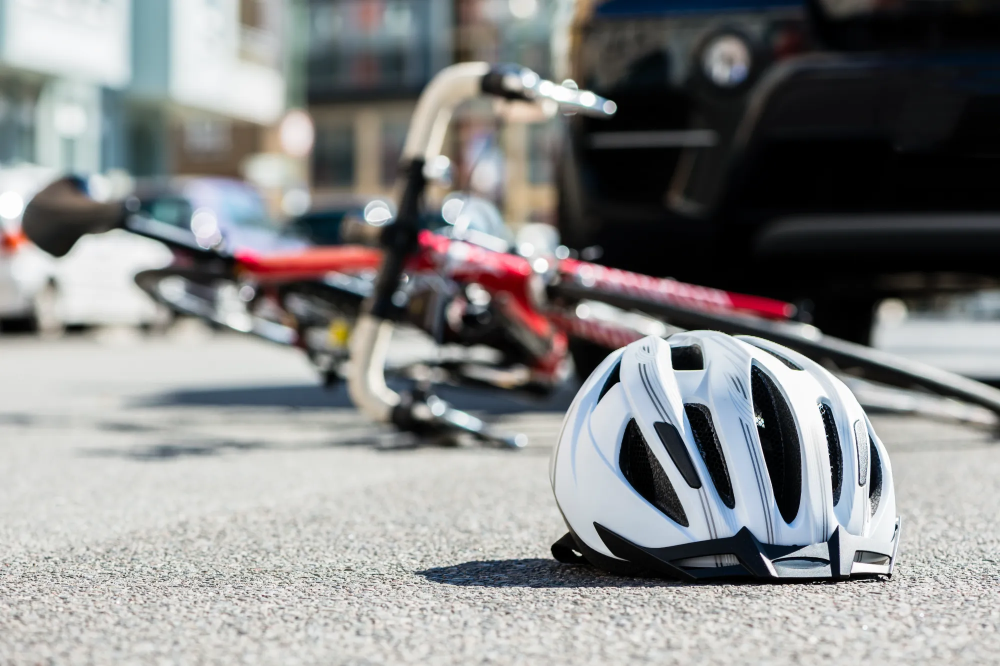 A white bicycle helmet rests on the pavement. A fallen red and black bicycle and the bumper of a dark car are visible in the background, suggesting a collision.