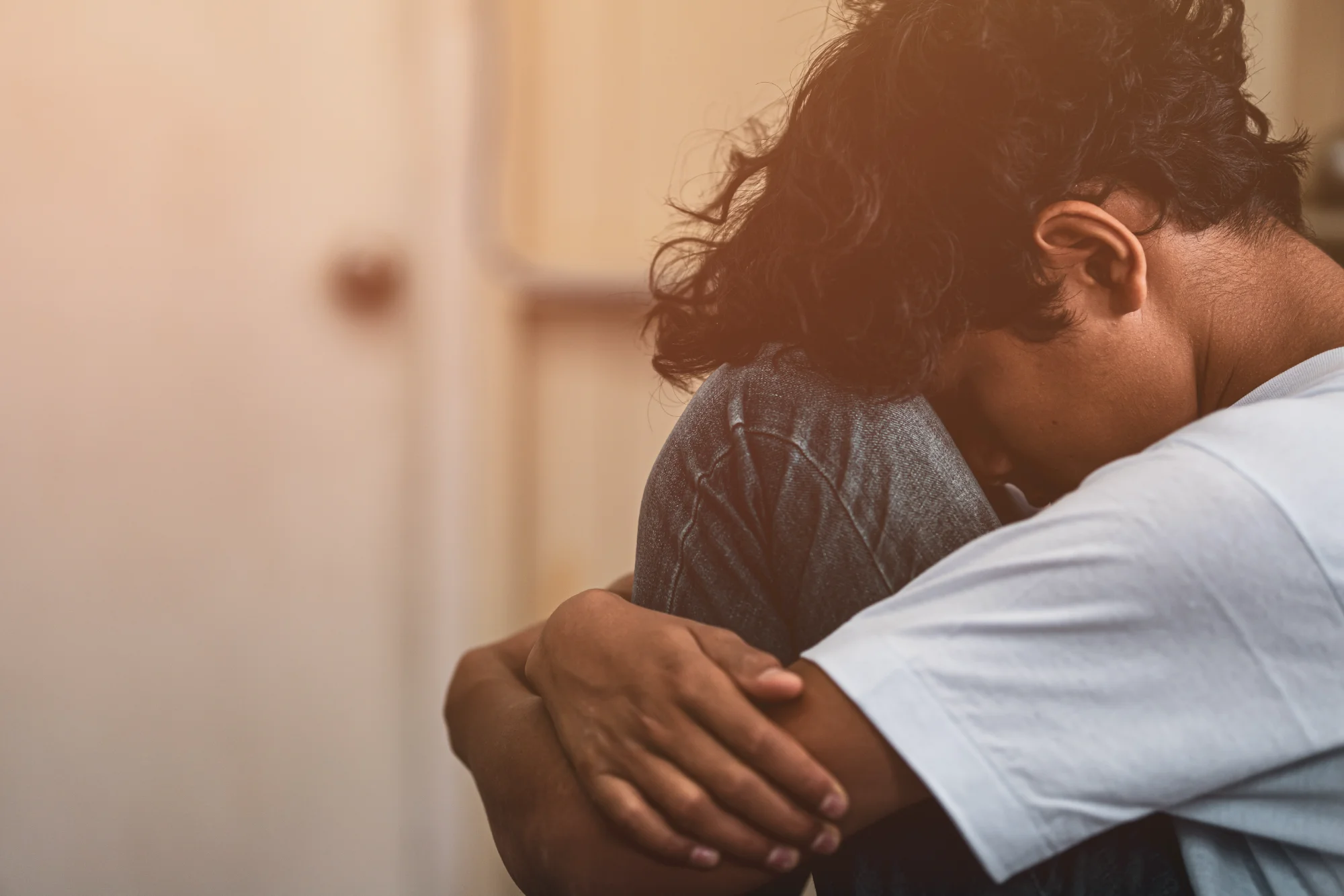 A person in a white t-shirt sits with their head bowed, clutching their knees tightly in a defensive or distressed posture.
