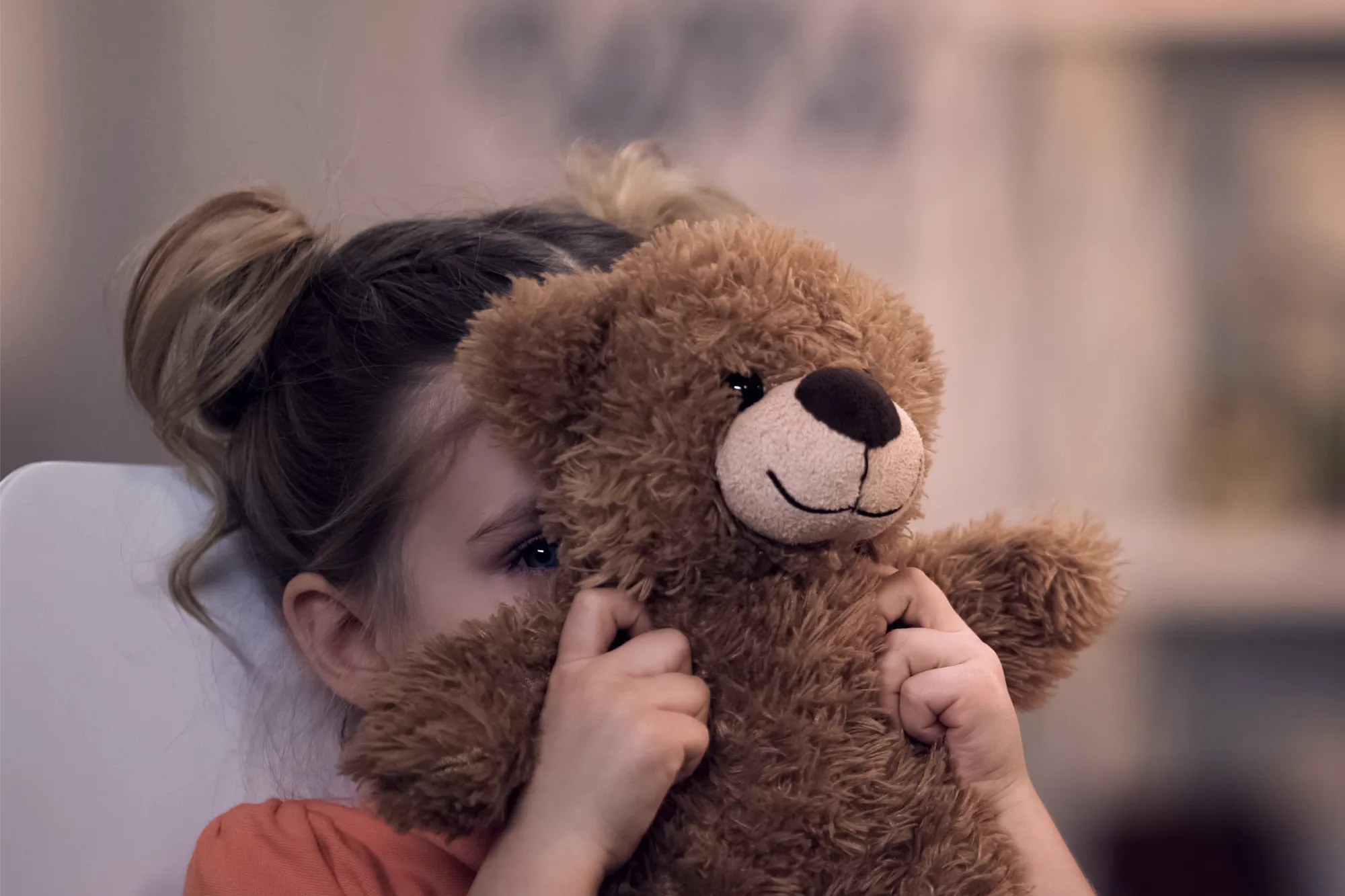 A child holds a large, brown teddy bear up to her face, hiding behind it with only one blue eye visible, giving a shy or playful expression.