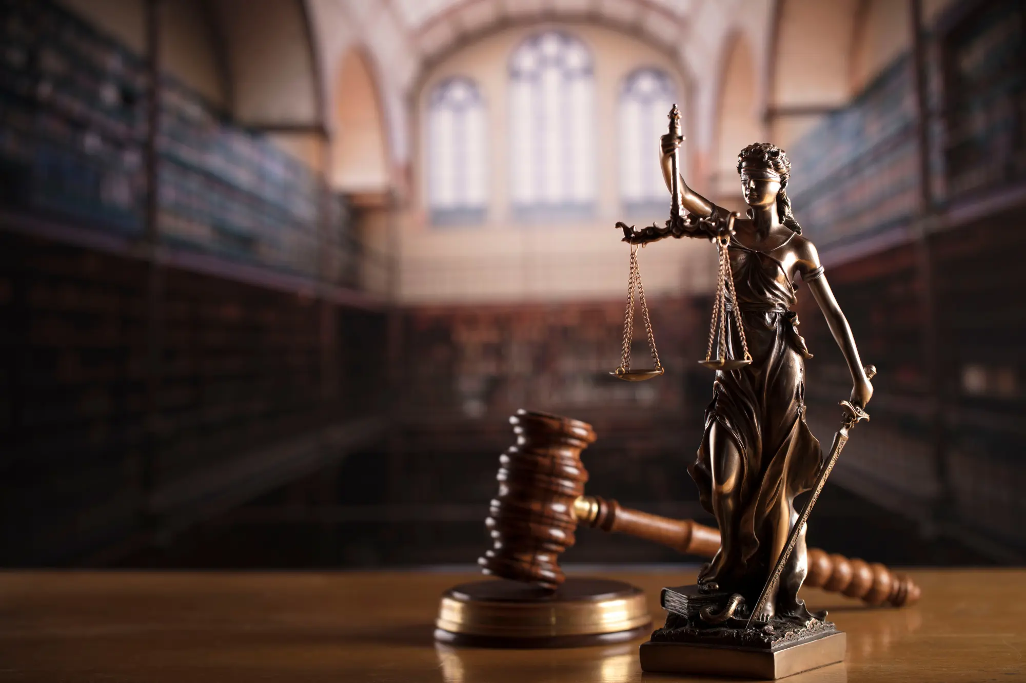 A close-up of a bronze statue of Lady Justice holding scales and a sword, positioned next to a gavel. The image is set in a dimly lit, historic law library.