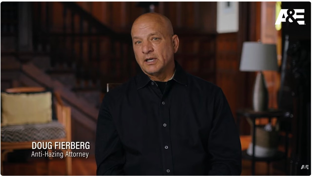 Anti-Hazing Attorney Doug Fierberg, bald and wearing a black collared shirt, sits for an interview in a dimly lit room with a dark wooden staircase.