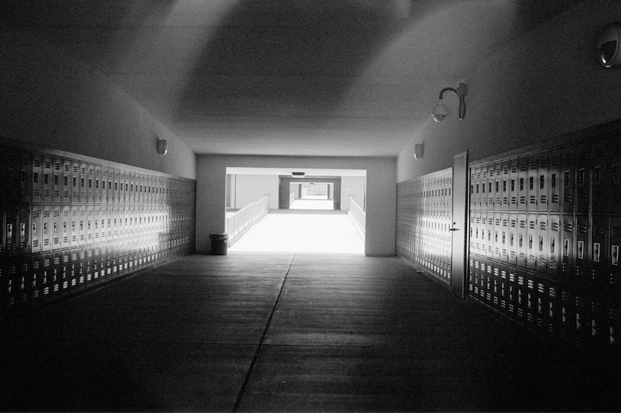 A black and white photo of a dark hallway lined with metal lockers, leading toward a bright, sunlit corridor.