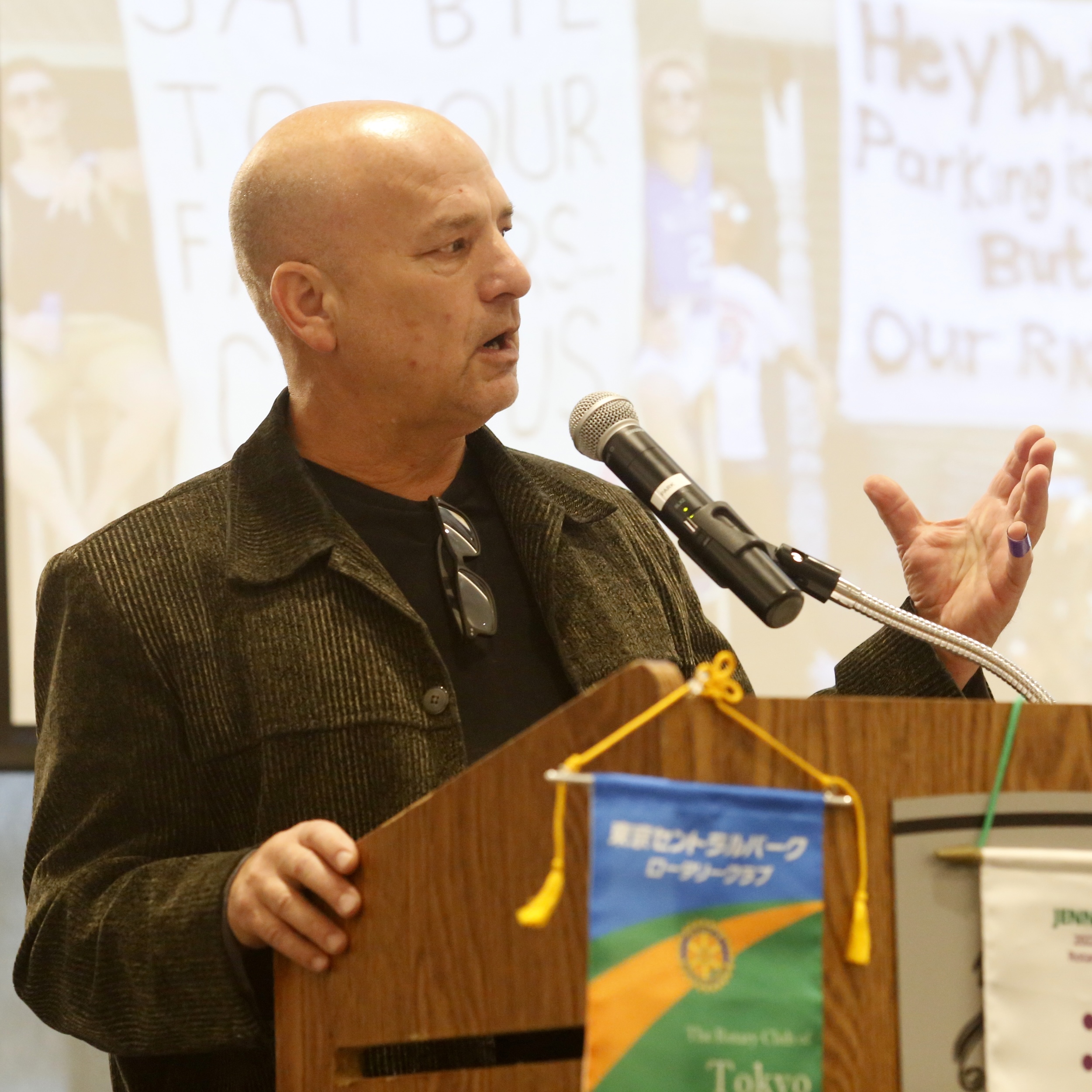 A bald man in a dark, textured jacket stands at a wooden podium, speaking into a microphone with a determined expression, gesturing with his right hand.