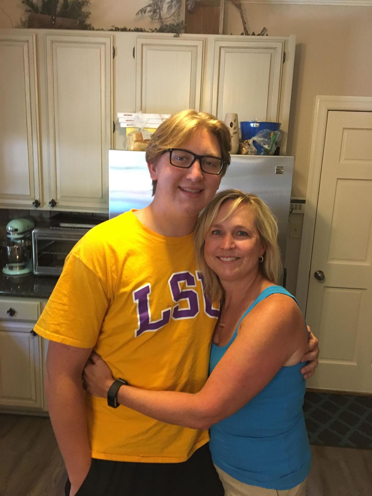 Max Gruver, a young man in an LSU t-shirt, stands next to his mother in a blue top, happily embracing in a kitchen setting. They both smile warmly.
