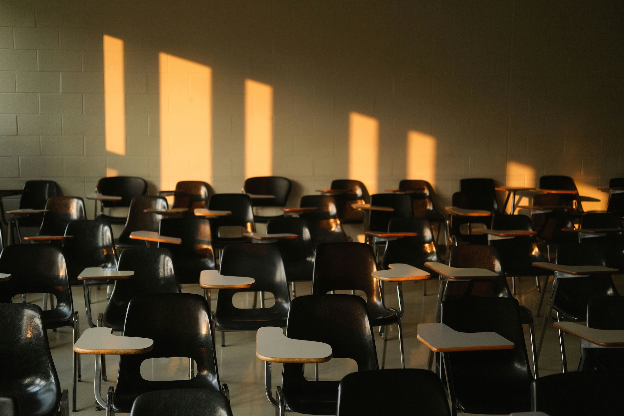Rows of black tablet arm chairs fill a dimly lit lecture hall. Warm sunlight casts long, vertical rectangles on the gray cinder block wall.