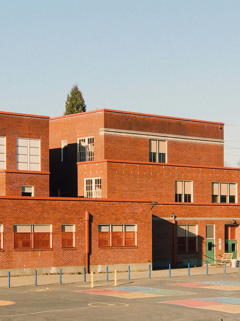 A large, tiered red brick school building stands under a clear sky, overlooking a paved playground area with faded colorful markings.