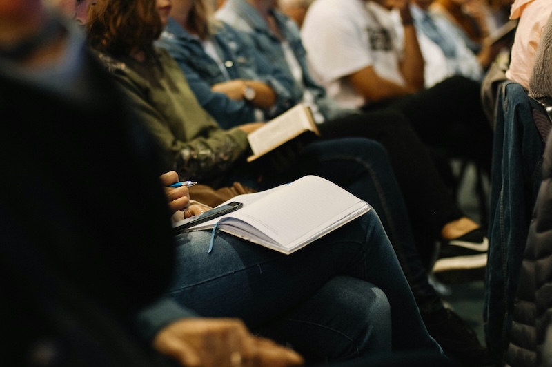 Students seated in a row, paying attention during a lecture. A person in the foreground holds a pen ready to take notes on an open notebook resting on their lap.