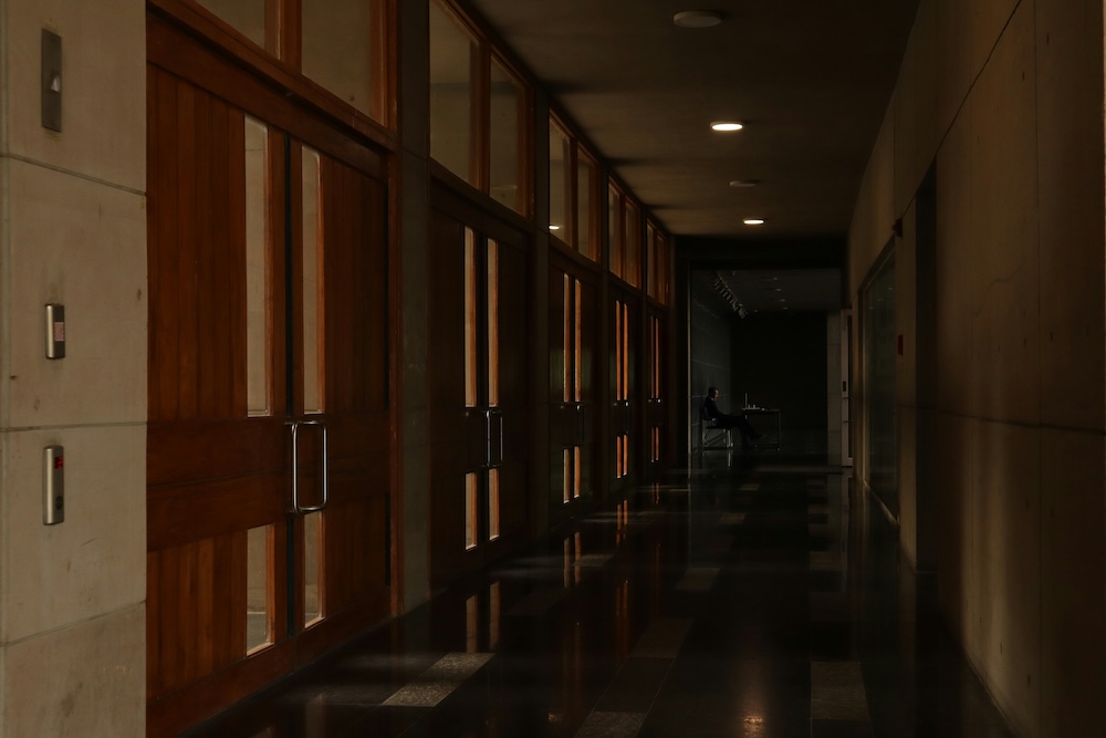 A long, dark office corridor with wooden paneled walls. A person is seated at a desk far down the hall, working alone in silhouette.
