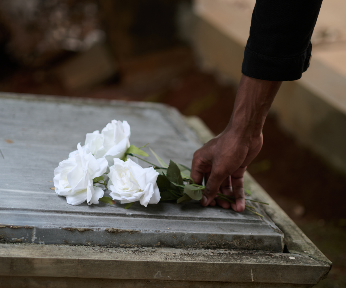 A hand in a black sleeve gently lays a small bouquet of white roses onto a weathered, gray stone grave marker.