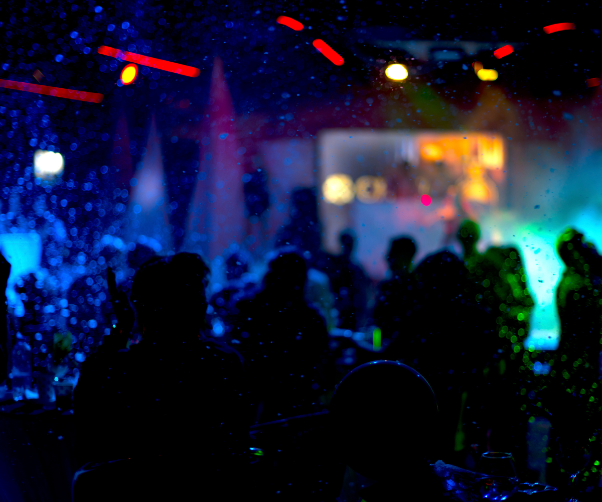 Silhouettes of a crowd of people at a lively indoor party, illuminated by dramatic blue and green stage lighting and colorful bokeh effects.