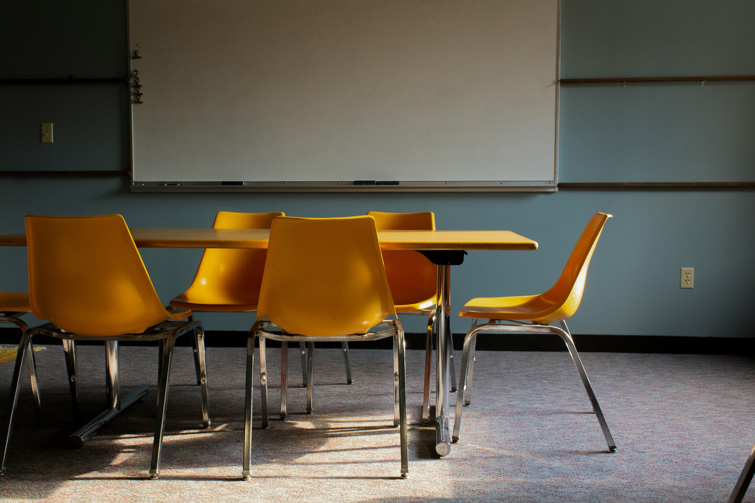 A table surrounded by four empty, bright yellow meeting chairs with chrome frames, set against a dusty blue wall and a large whiteboard.