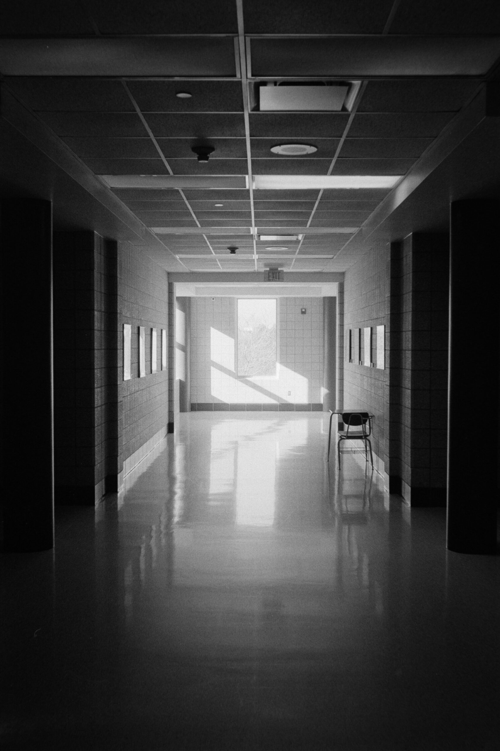 A high-contrast black-and-white photo of an empty institutional hallway with a polished floor reflecting bright light from a far window. A single chair sits near the wall.