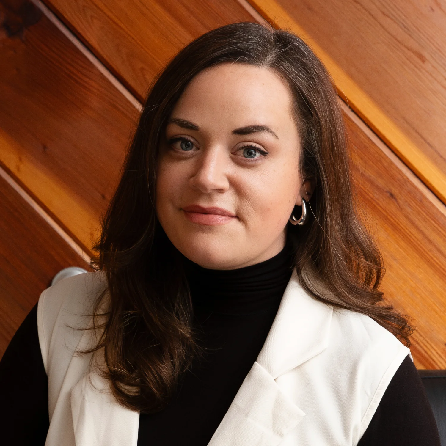 Bevin Buchler smiles professionally in a white vest and black top, standing in front of a warm, diagonally oriented wooden wall.