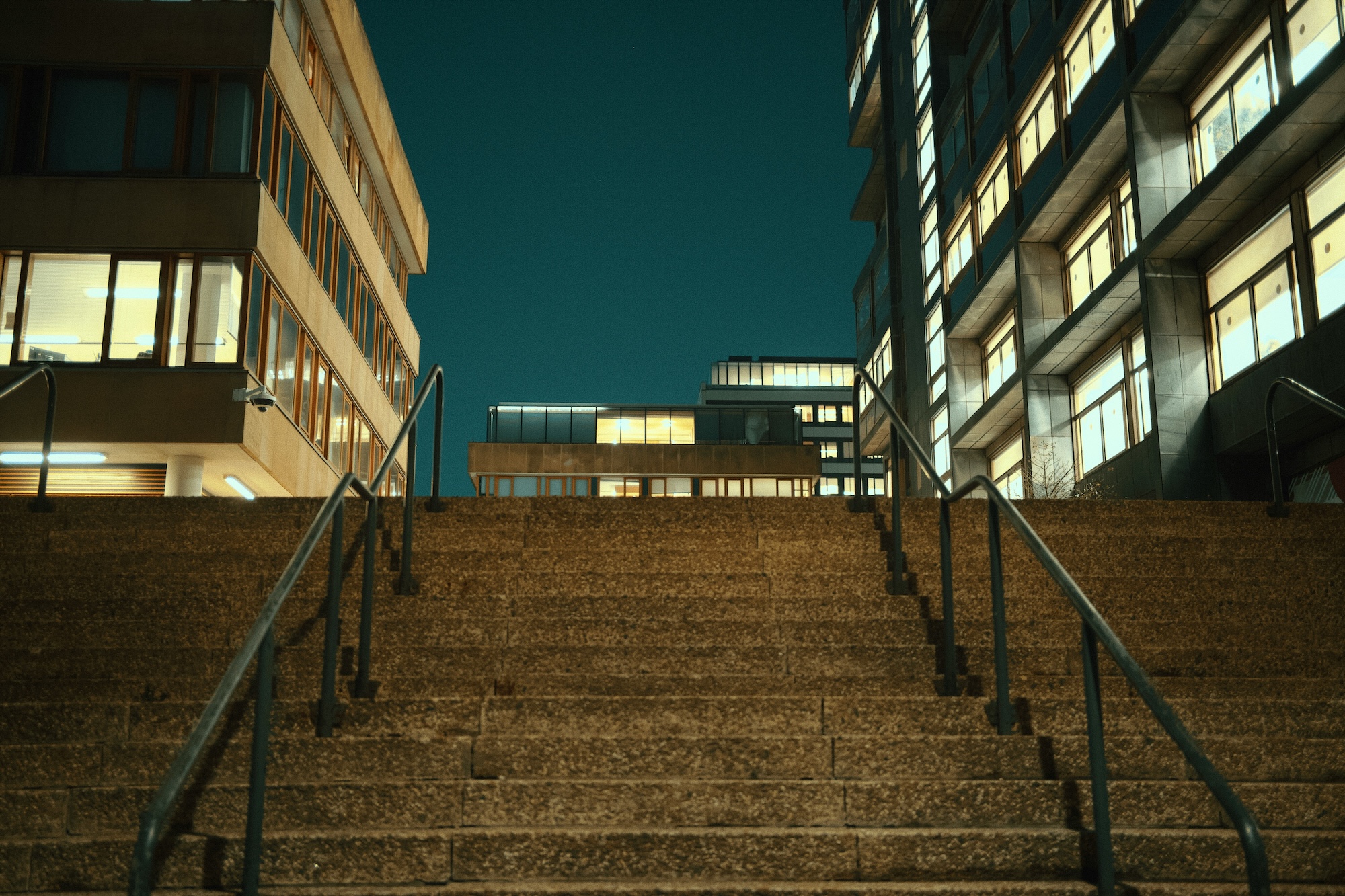 A wide stone staircase leads up between two large campus buildings whose many windows are brightly illuminated against a dark night sky.