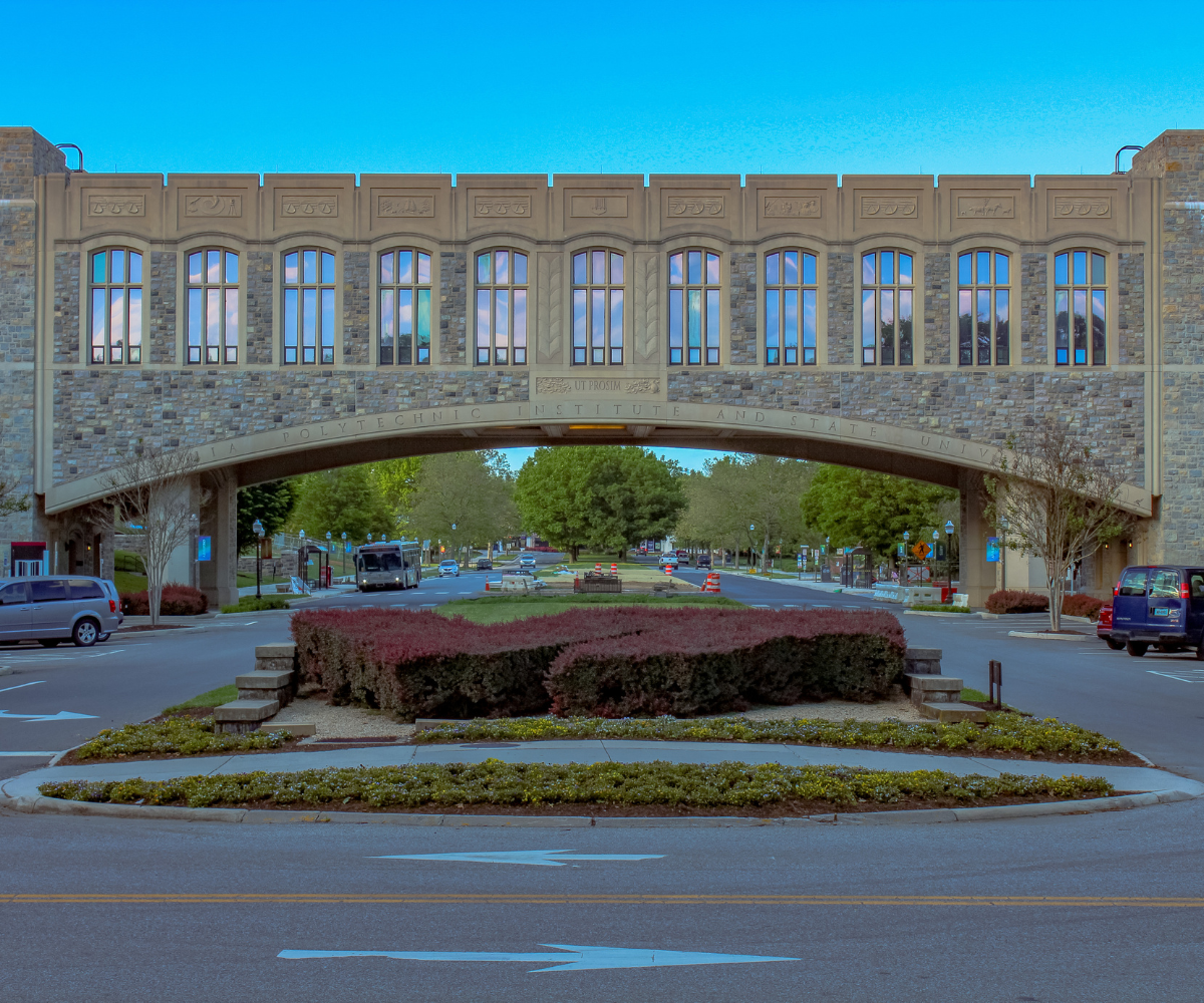 A massive stone bridge with arched windows crosses a street at Virginia Tech, framed by a traffic circle with red and green landscaping.