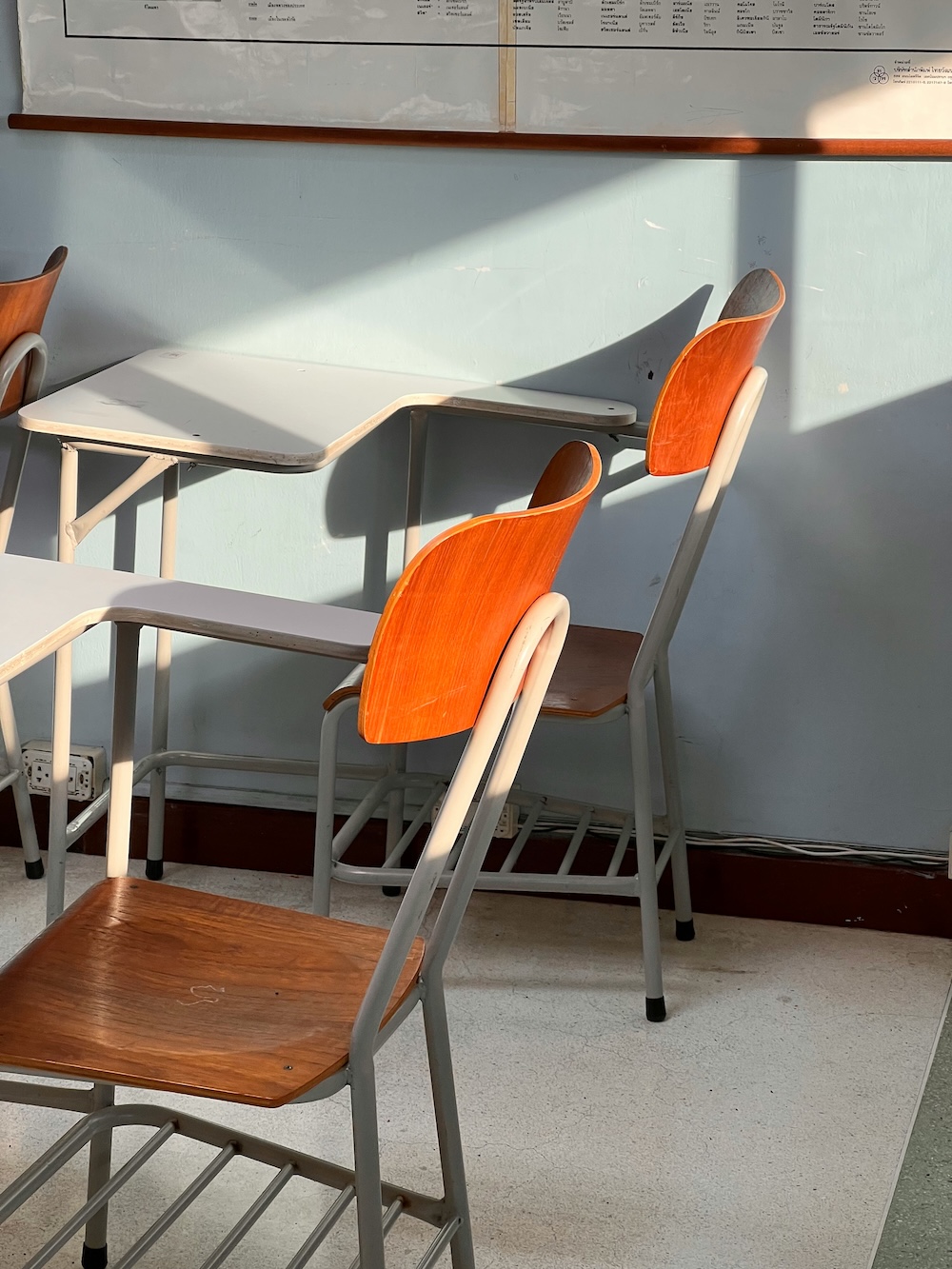 Sunlight casts strong shadows across a light blue classroom wall behind two unoccupied student chairs with curved, bright orange backs.