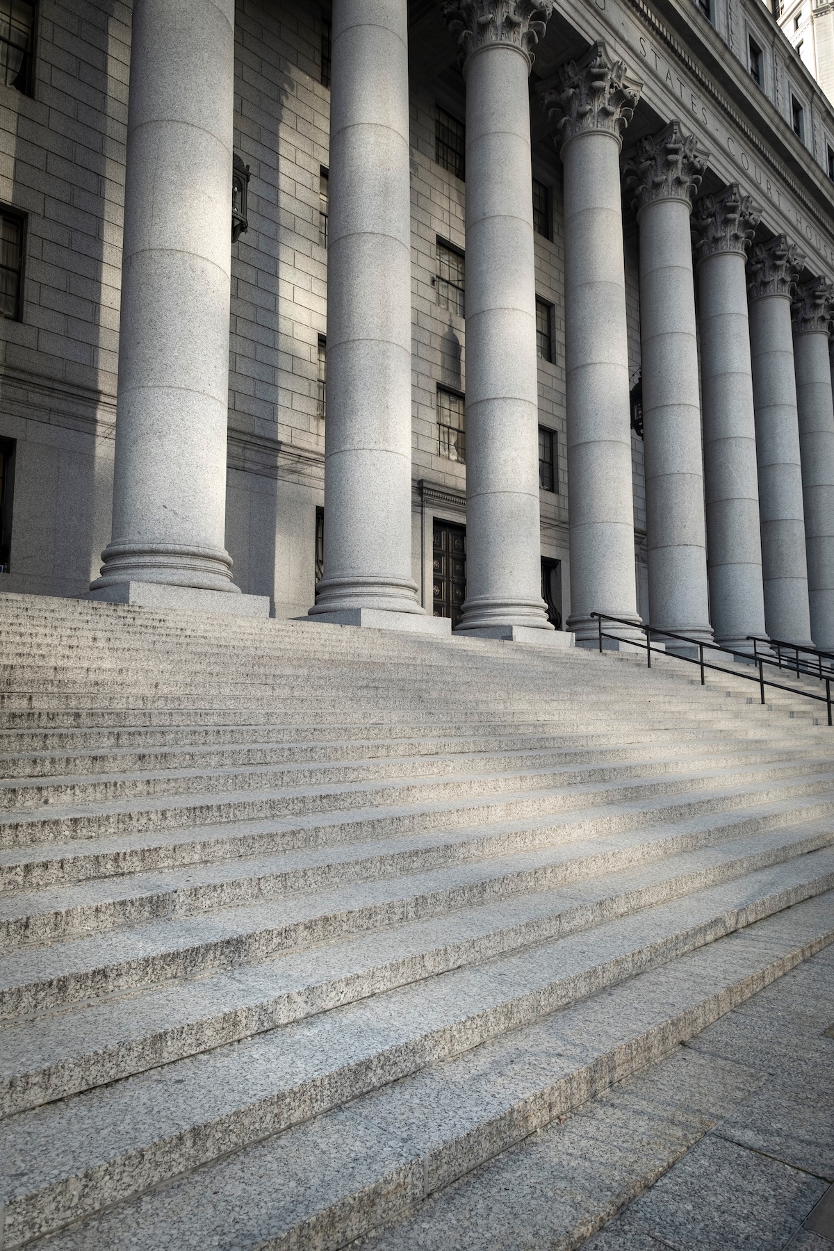 A grand entrance to a stone courthouse featuring wide granite steps leading up to a massive colonnade of classical columns with ornate capitals.