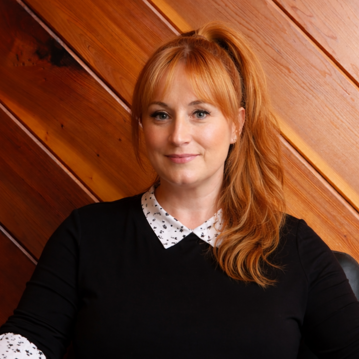Erika Klockziem sits in a black chair against a diagonal wood-paneled wall. She wears a black top with star-patterned white accents and looks forward with a calm smile.