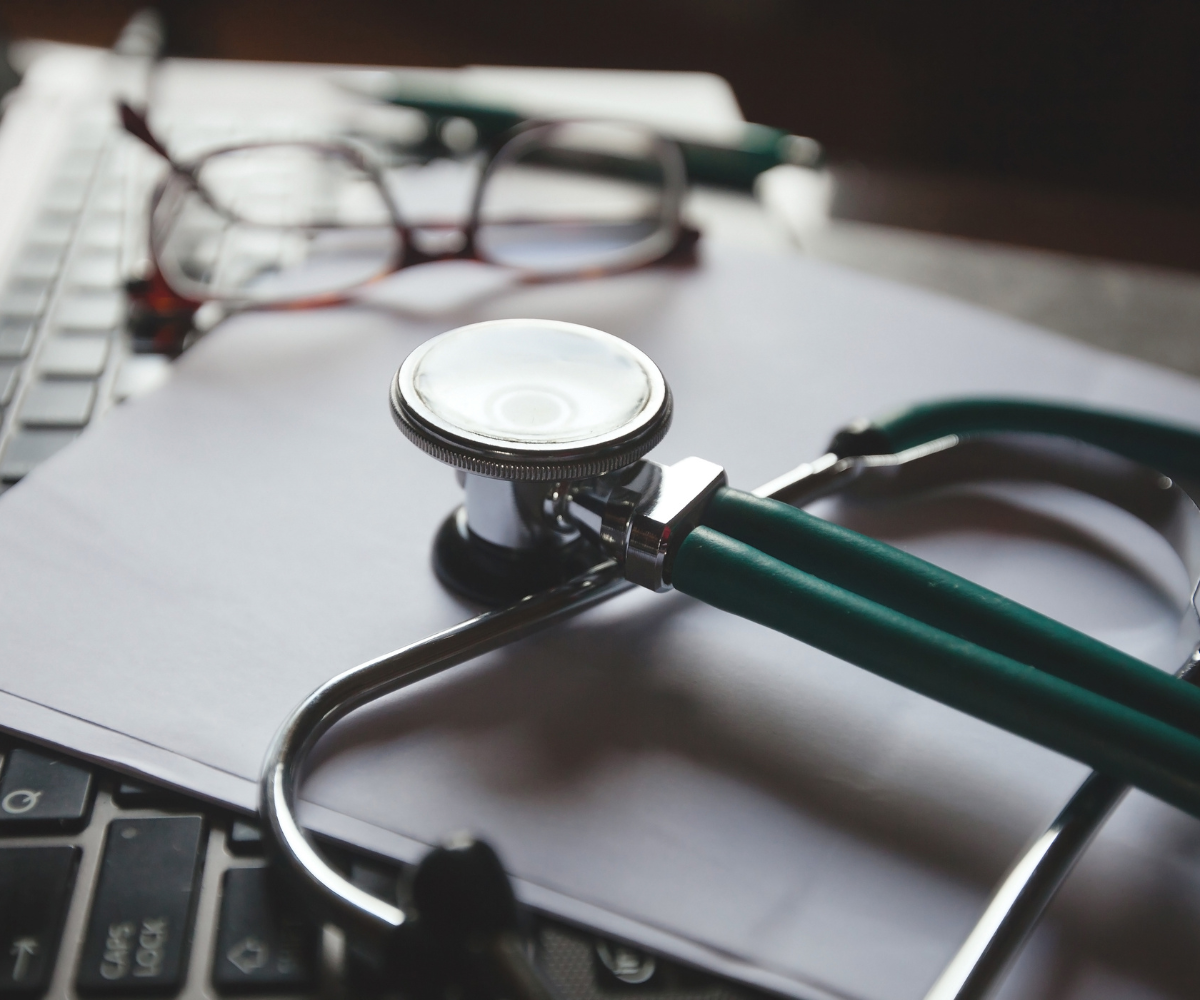 A green stethoscope, eyeglasses, and a pen rest on a white document over a laptop keyboard, representing a detailed review of medical documentation.