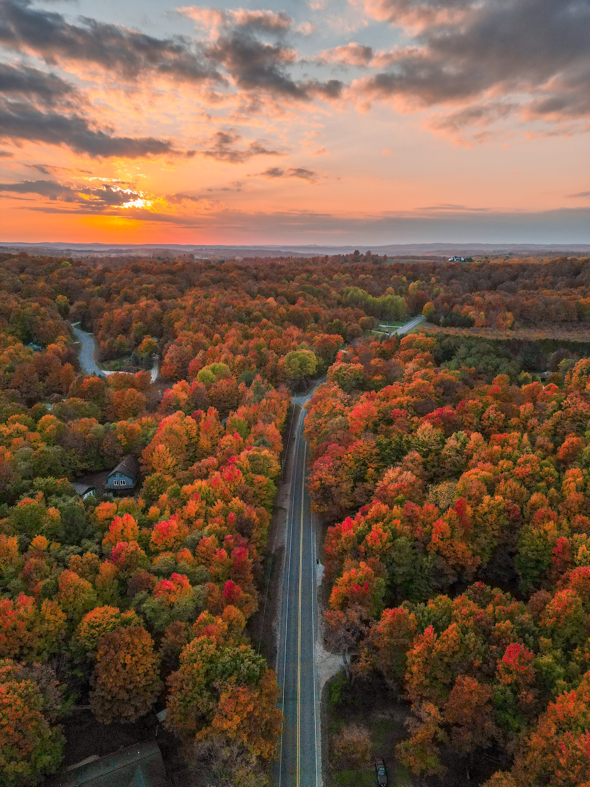 An aerial view of an empty road in Northern Michigan lined with brilliant red and orange fall foliage under a dramatic sunset.