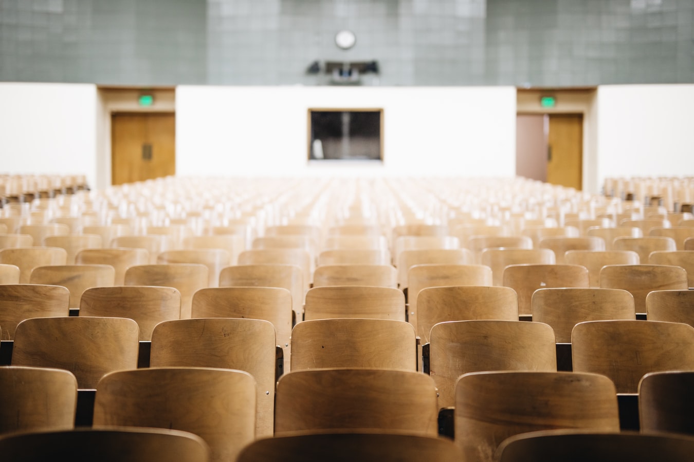 A large, empty lecture hall is seen from the back, showing endless, tightly packed rows of light wooden seats leading toward a white wall with doors.