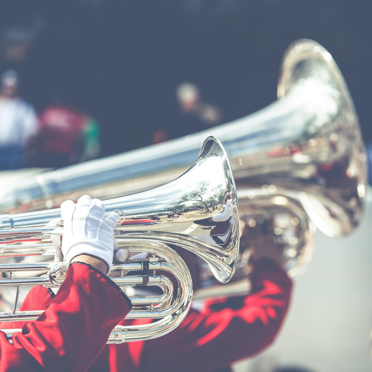 A marching band member in a red uniform jacket and white gloves plays a shiny silver tuba during a performance.
