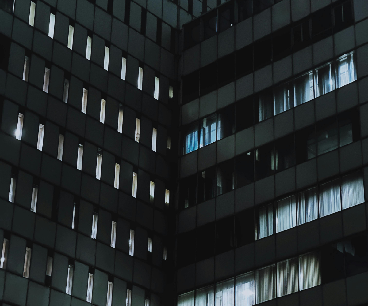 The exterior of a tall modern building at night. Several interior office lights glow faintly through the long windows and curtained panes.
