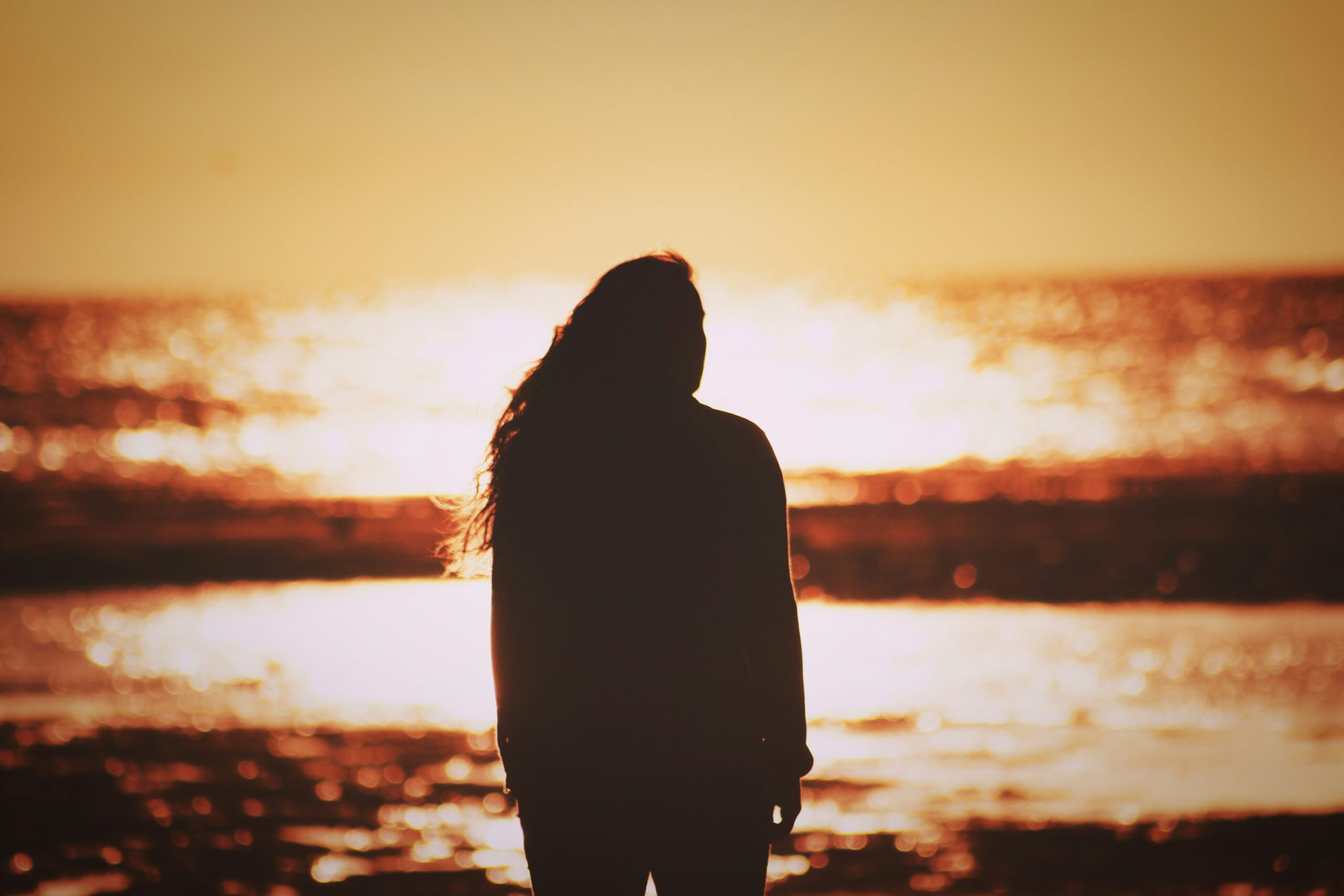A silhouette of a person with long hair stands facing the ocean, backlit by a brilliant orange and yellow sunset reflecting off the water.